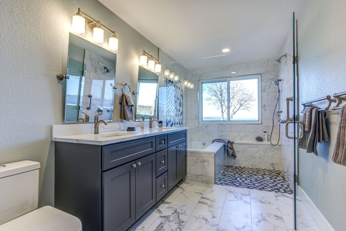 Modern bathroom with a dark gray double vanity, marble-style flooring, a large soaking tub, and a glass shower stall.
