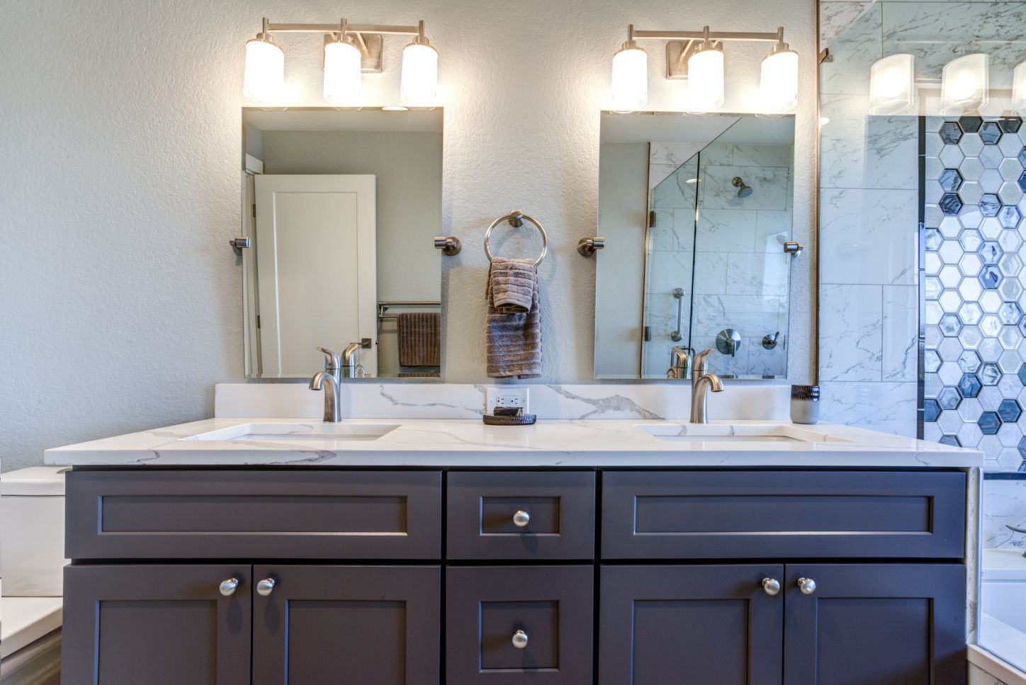 A double-sink bathroom vanity with dark gray cabinets, white marble countertops, two mirrors, and overhead lighting.