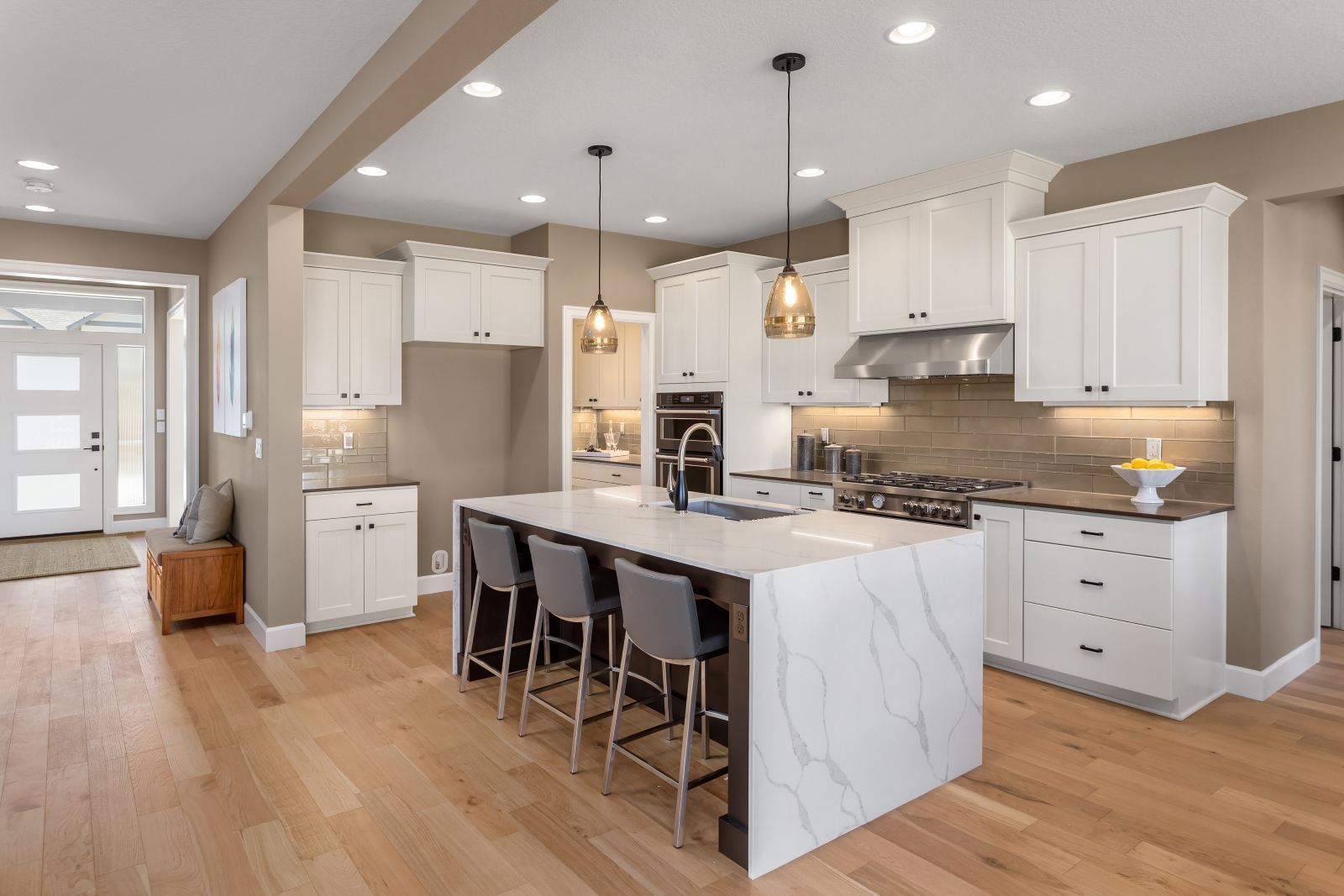Modern kitchen with a white marble island, grey bar stools, white cabinetry, and light wood floors.
