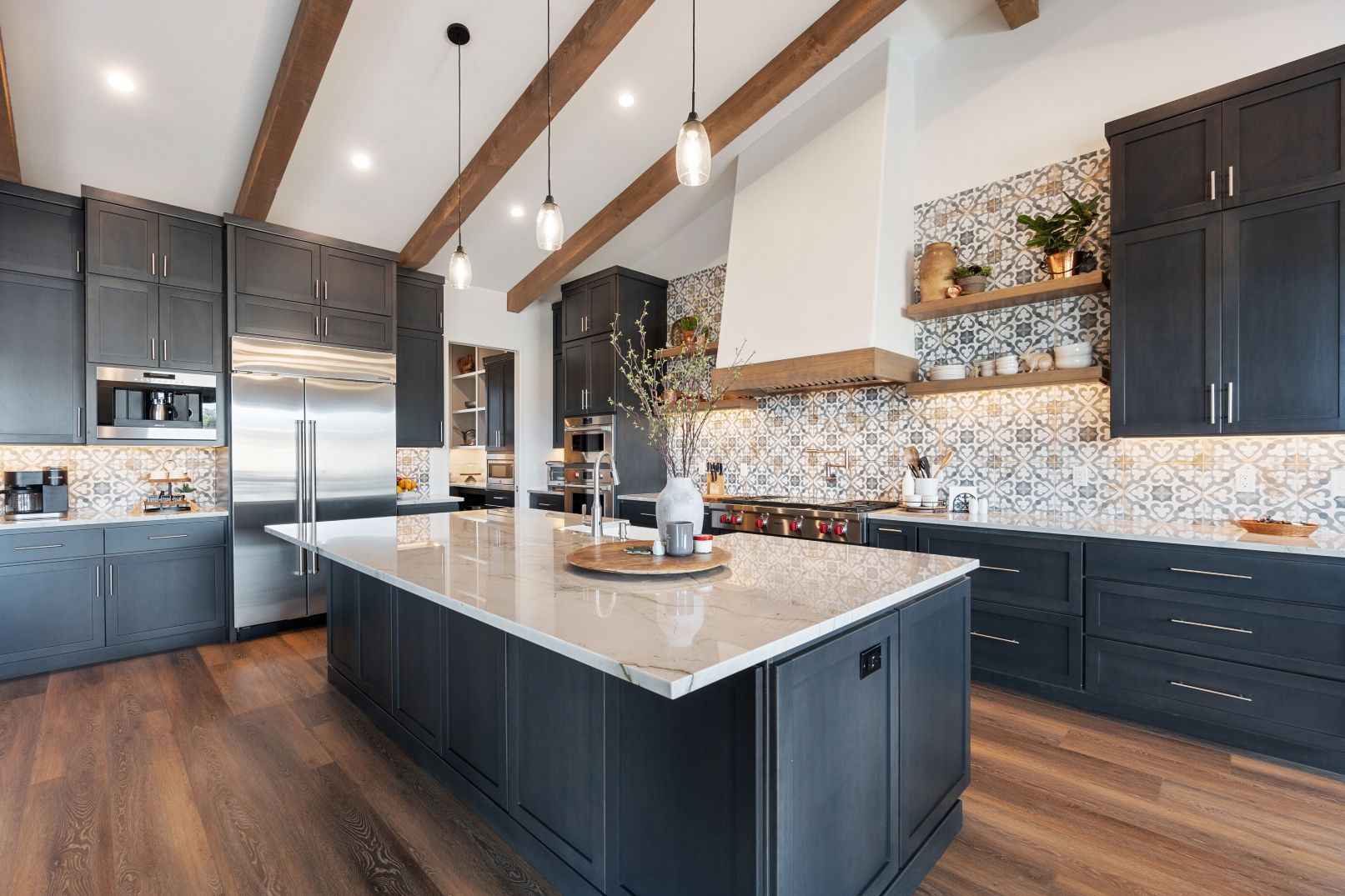 A modern kitchen with dark cabinets, a large granite island, patterned tile backsplash, and wooden ceiling beams.