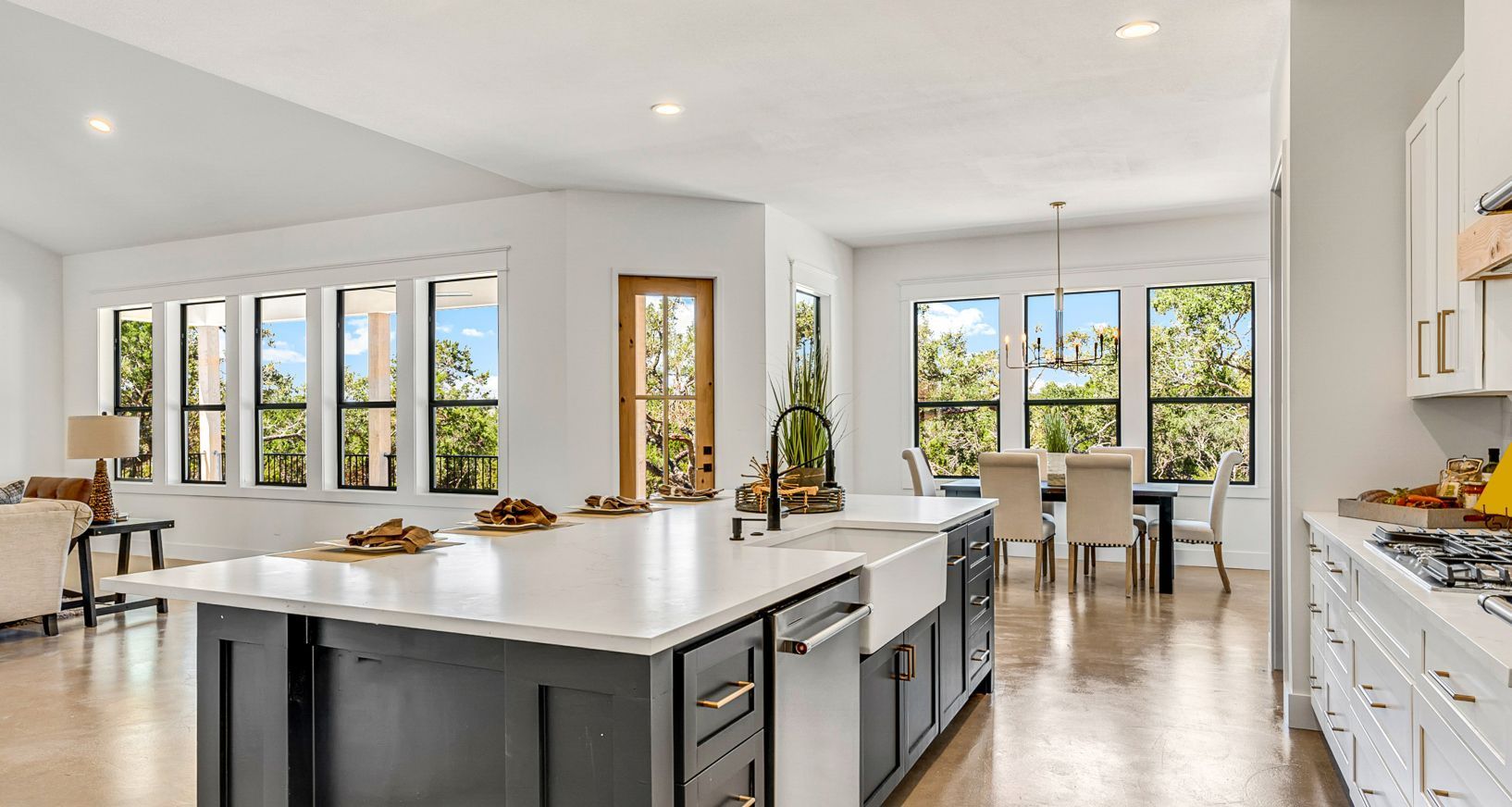 A modern, open-concept kitchen with a large dark gray island, white cabinets, and a dining area with views of greenery.