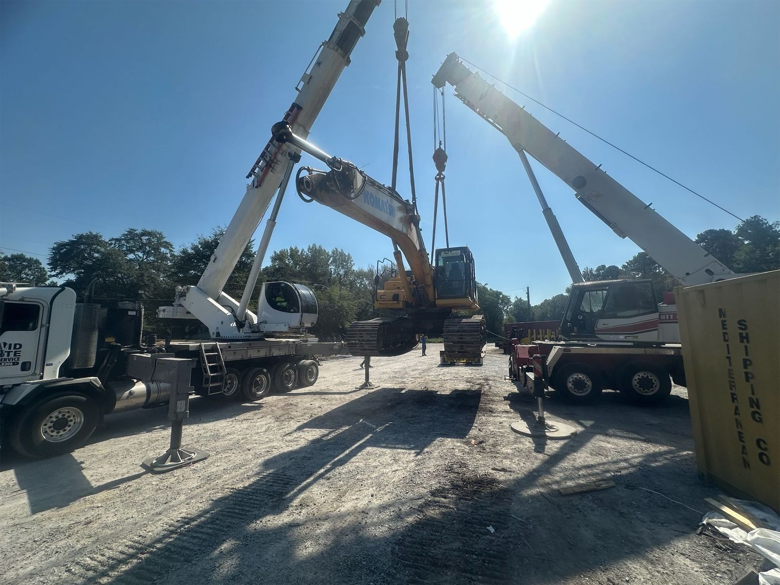 Two cranes lift an excavator between trucks on a construction site under a sunny, blue sky.