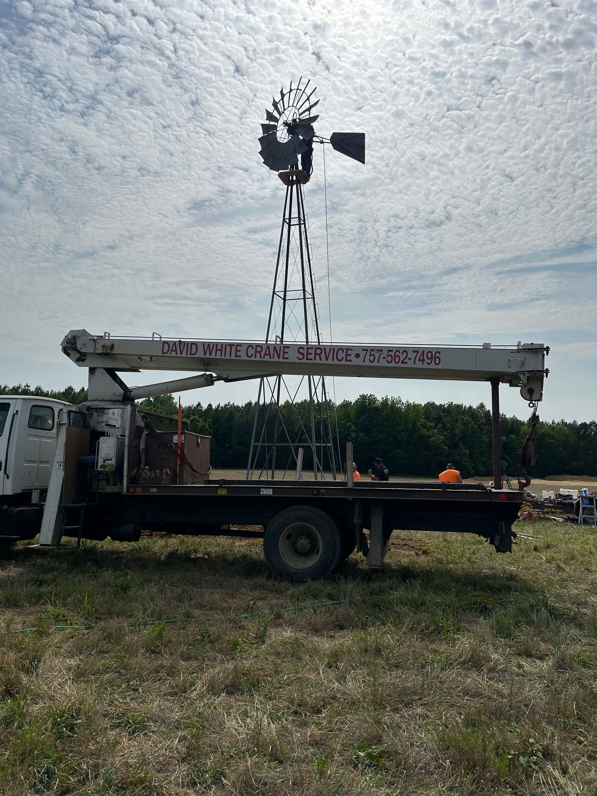 Flatbed truck with a large metal tower and wind turbine-like rotor in a grassy field under a cloudy sky