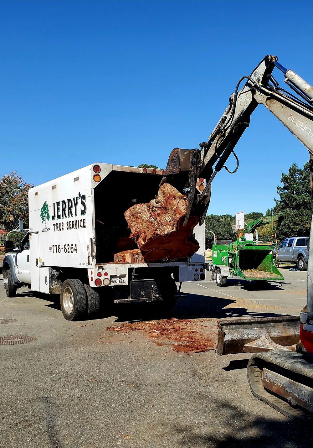 An excavator loading tree debris into a white truck from Jerry's Tree Service. A wood chipper sits nearby on a paved area.