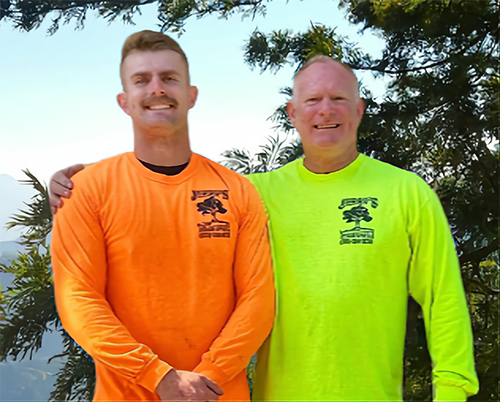 Two men in long-sleeve shirts with a tree logo, standing outdoors. One in orange, one in yellow, smiling, arm around the other.
