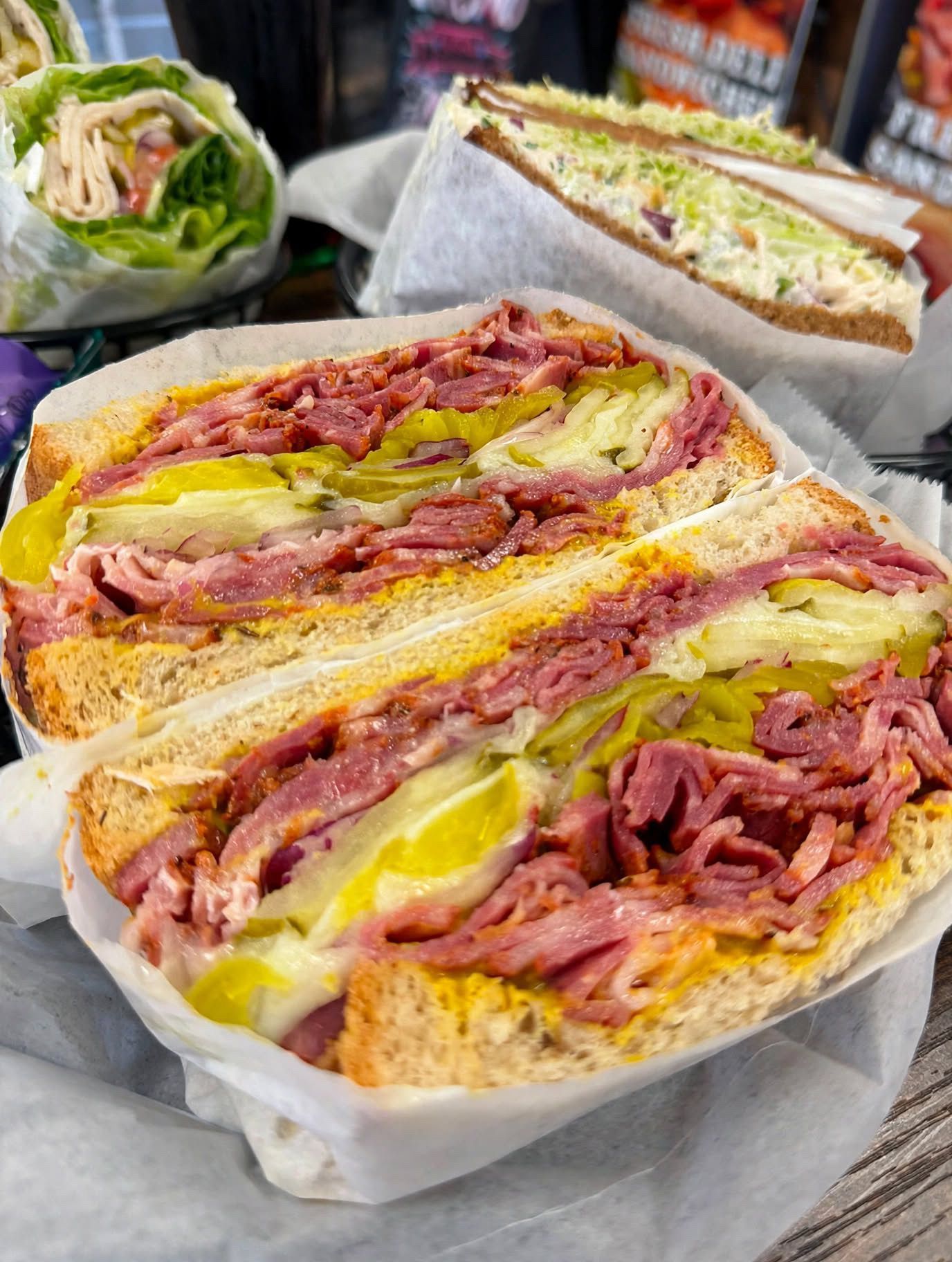 Close-up of a deli sandwich stuffed with pastrami, pickles, and mustard in wax paper.