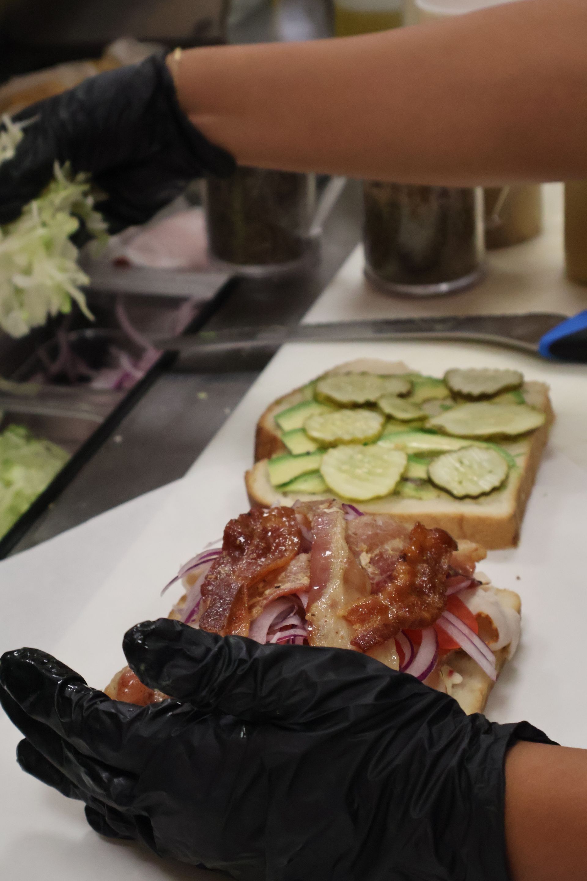 Gloved hands preparing a sandwich with sliced cucumbers and bacon on a white plate in a kitchen.
