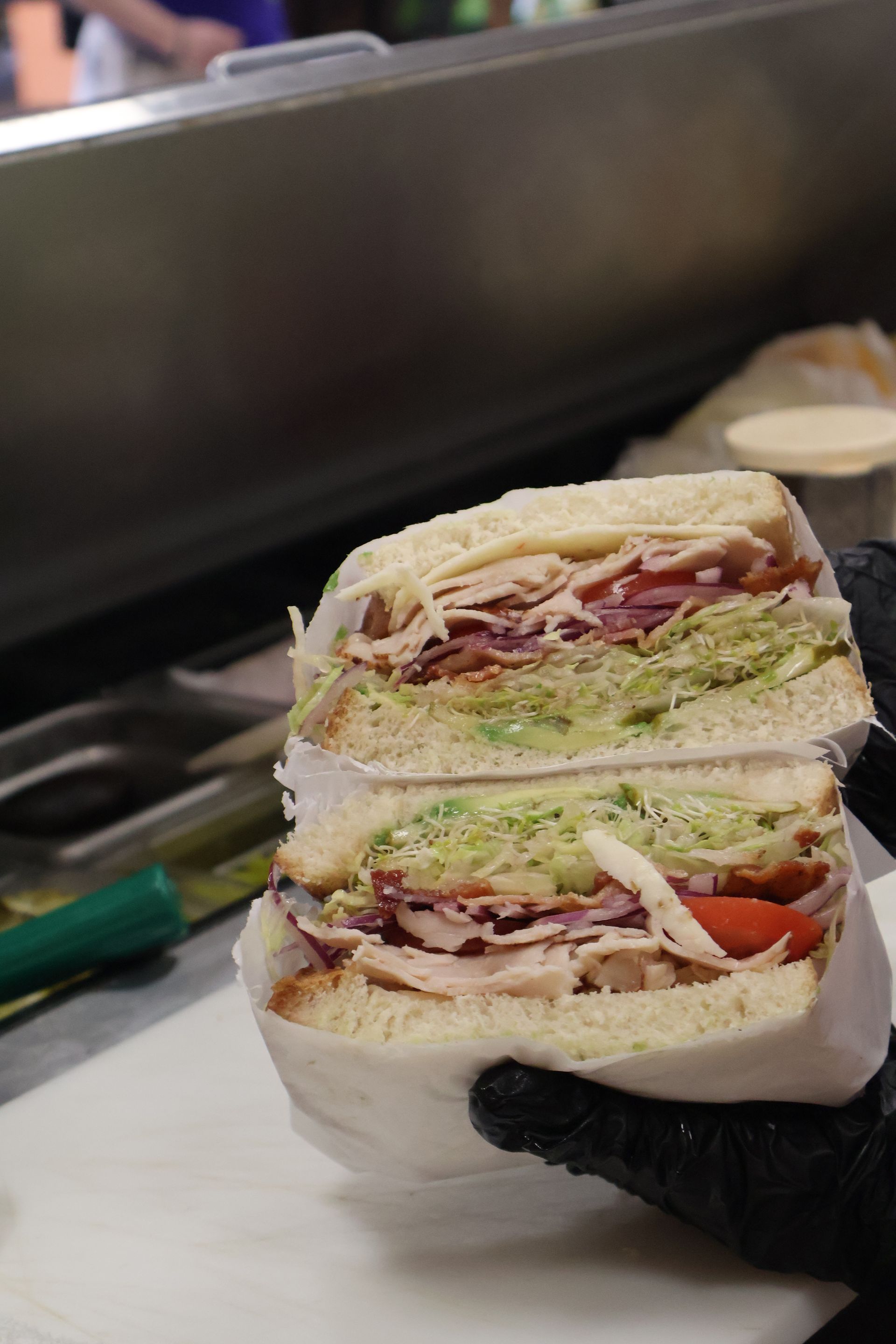 A hand in a black glove holds a stacked sandwich with lettuce, tomato, turkey, and bread layers.