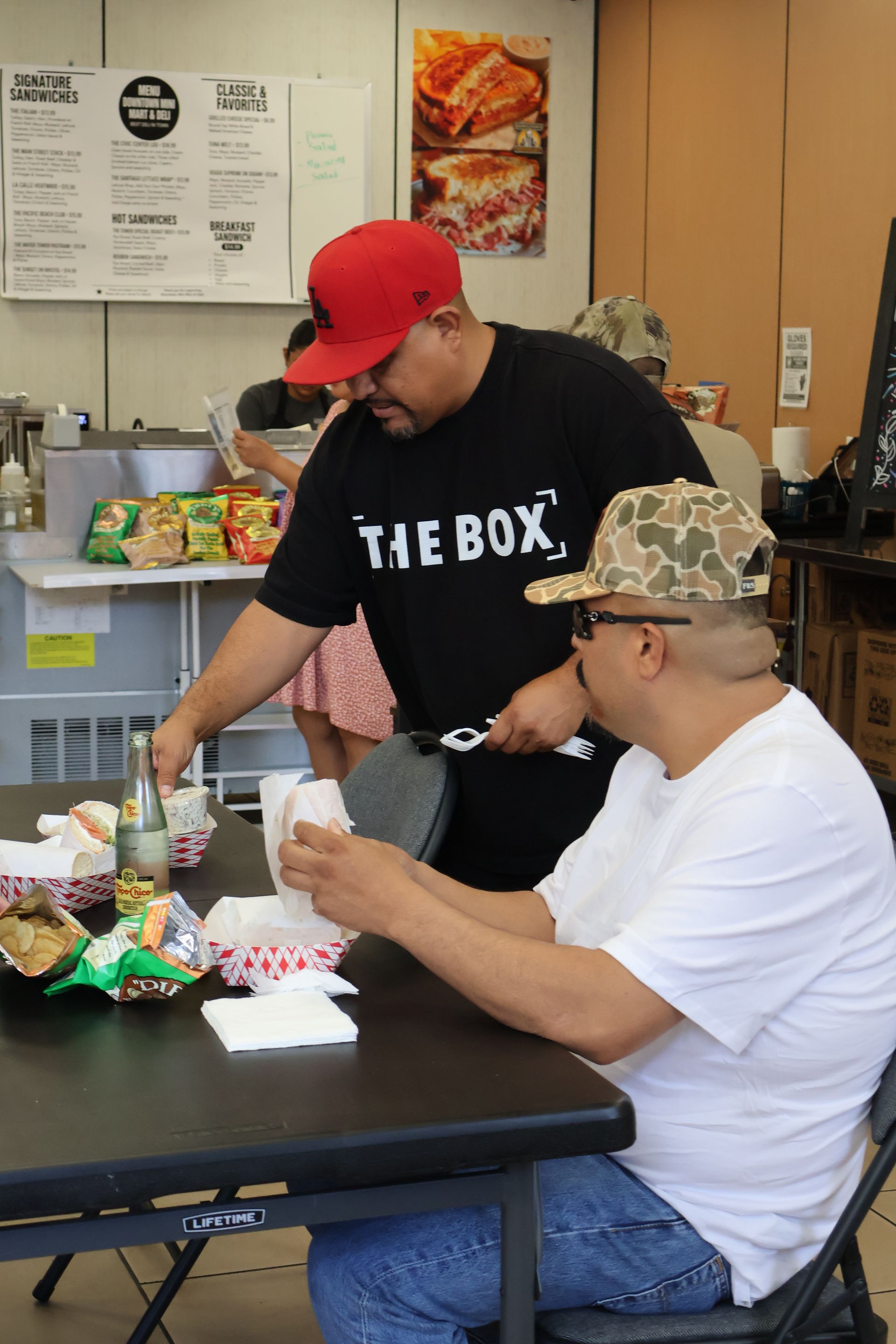 People at a table in a casual restaurant, one standing and arranging food while the other sits in a camo cap.