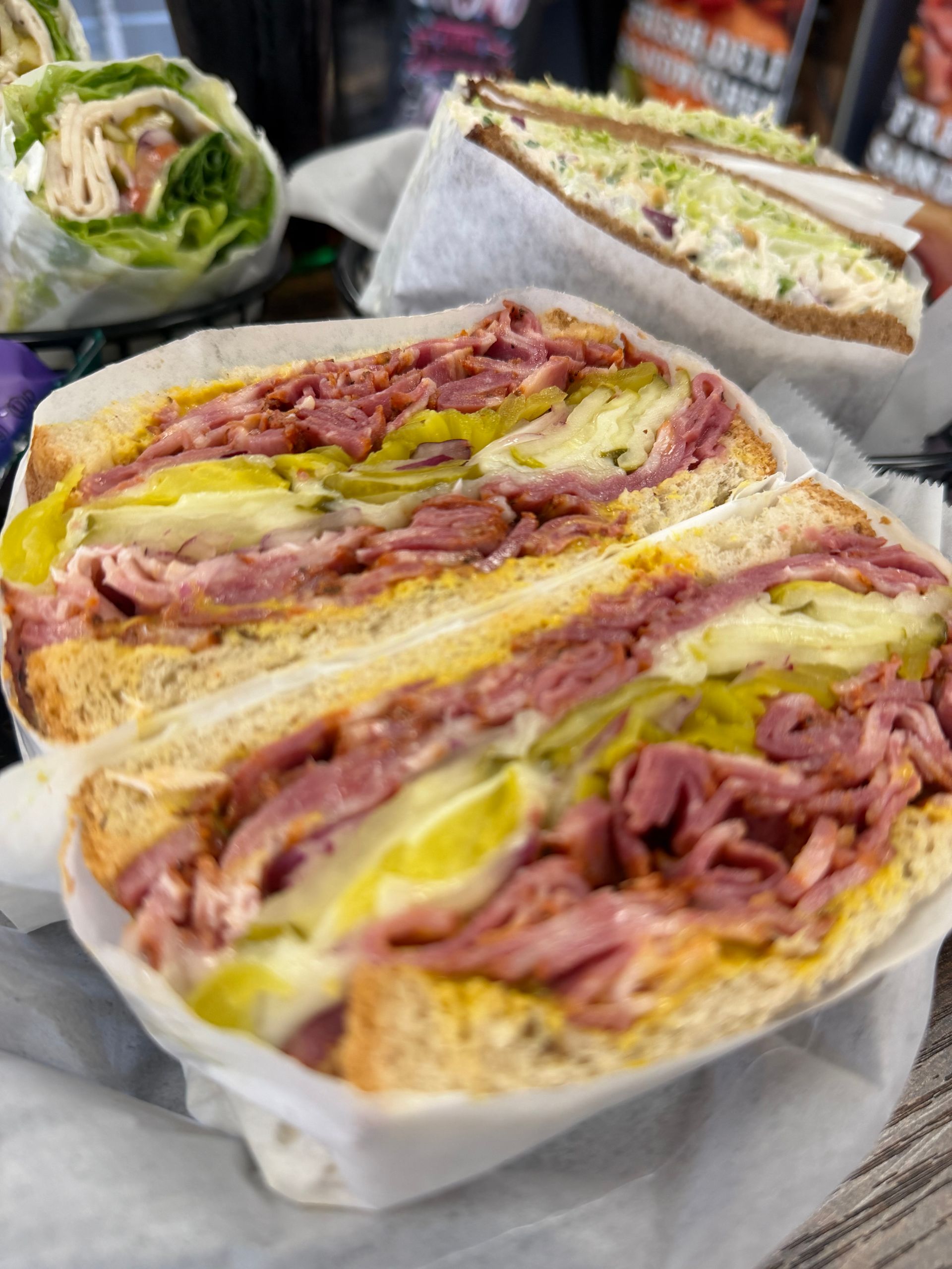 Stacked deli sandwiches with ham, cheese, and pickles on white paper wrappers.