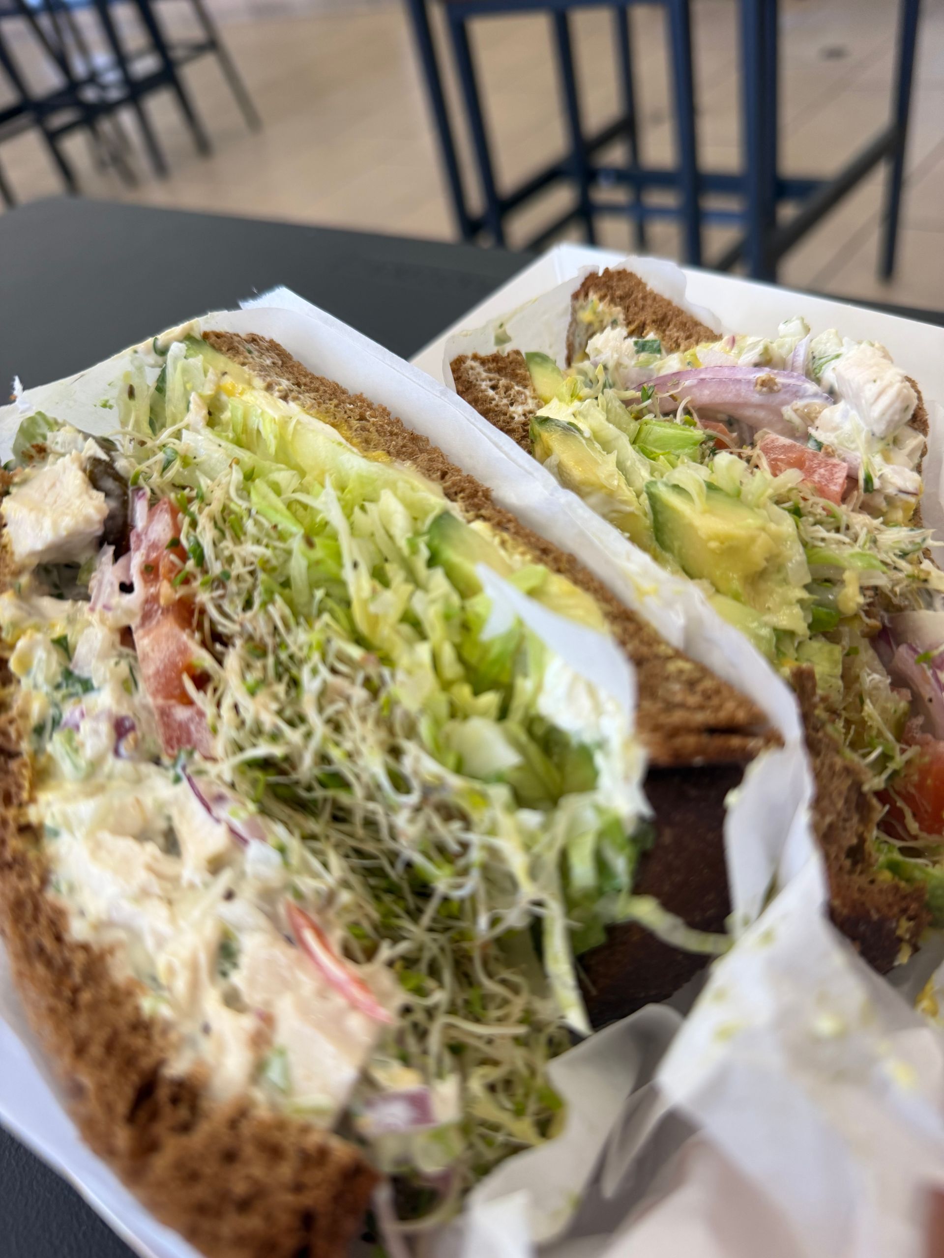 Two half sandwiches with shredded lettuce and fillings on a tray in a café setting.