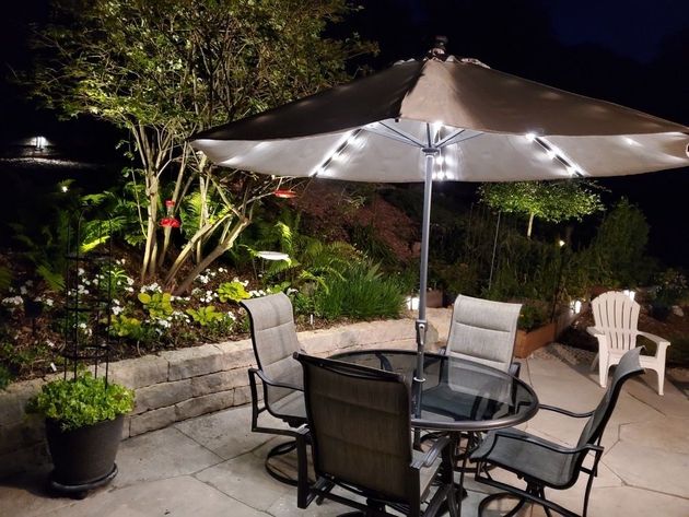 Patio seating under a lit umbrella beside a landscaped garden at night