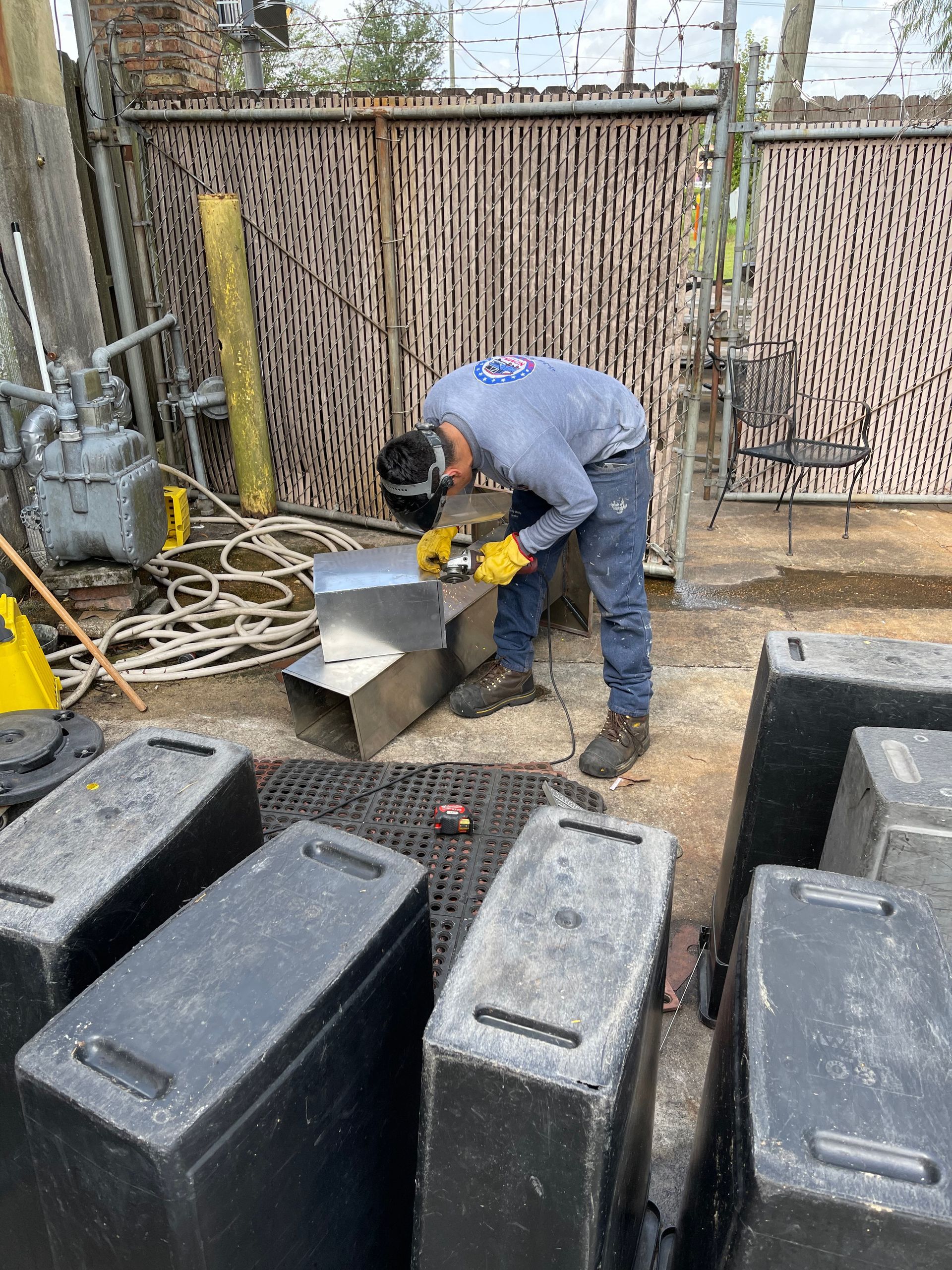 Man wearing safety glasses welding a metal box outdoors, surrounded by black containers.