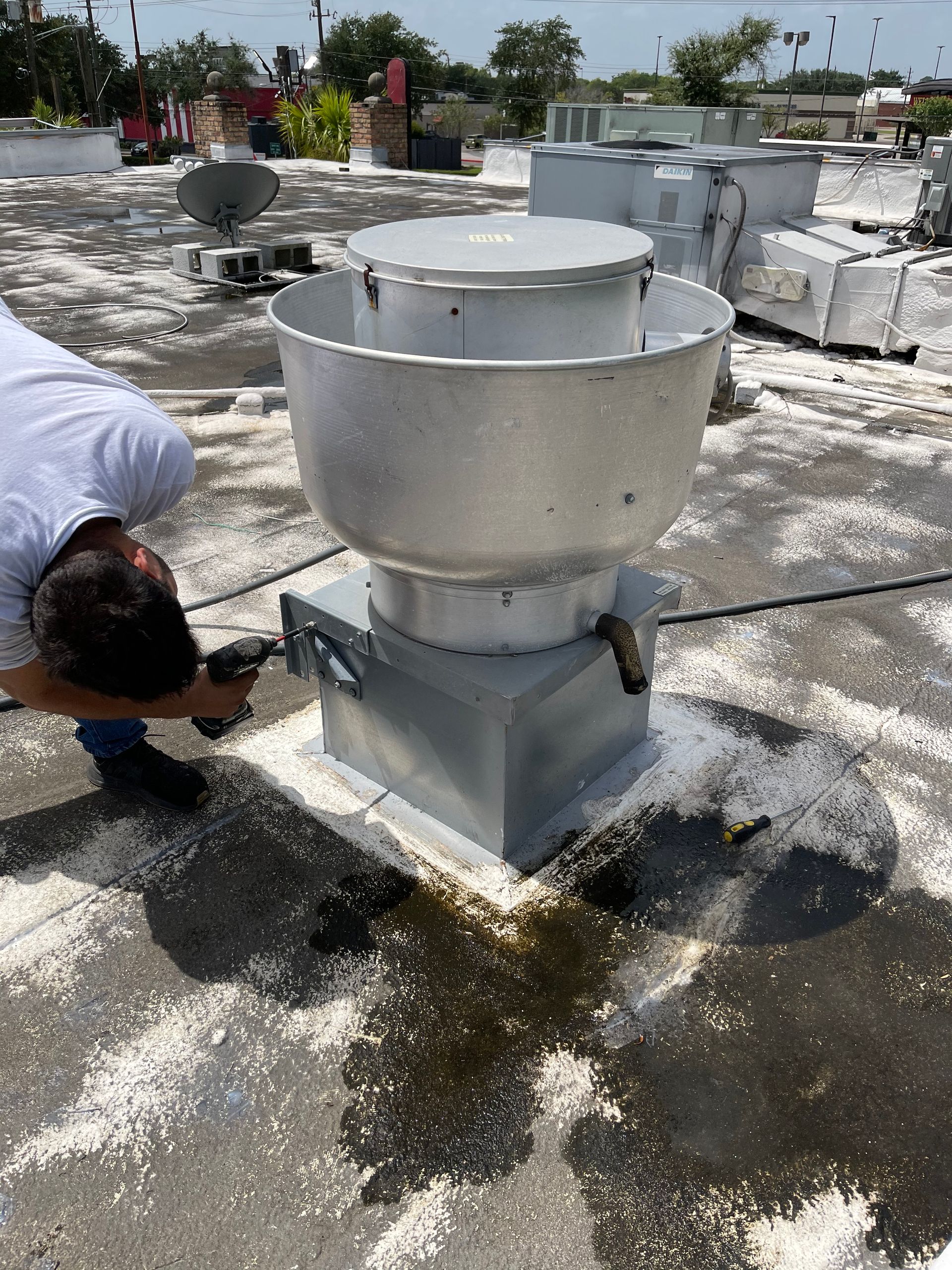 A man examines a large, silver rooftop vent on a light-colored roof with debris.
