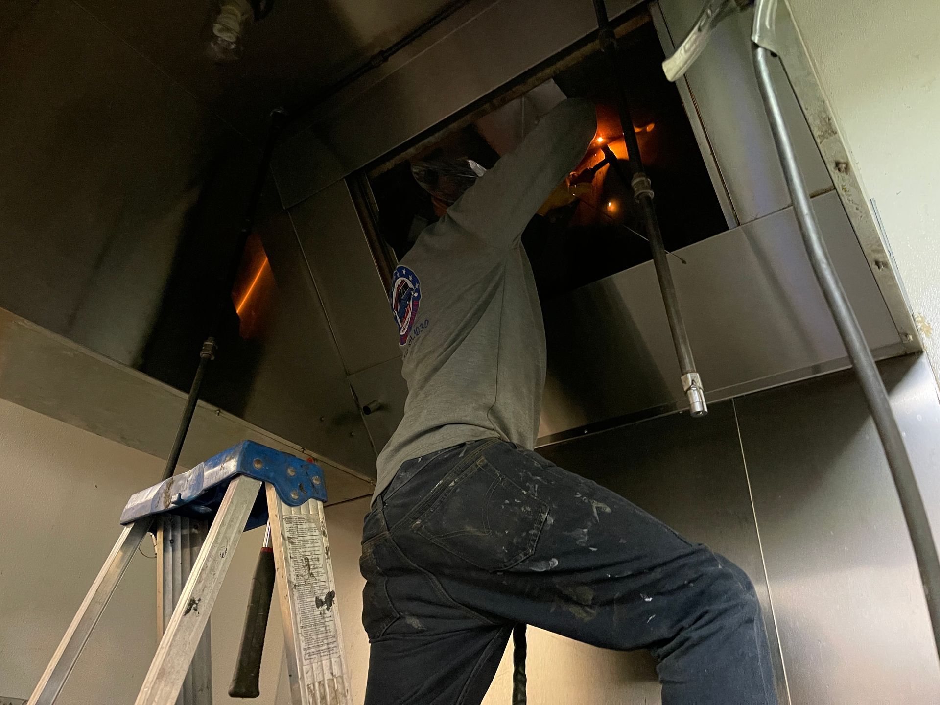Person on ladder, repairing kitchen exhaust hood. Inside, gray sweatshirt, black jeans, working with tools.