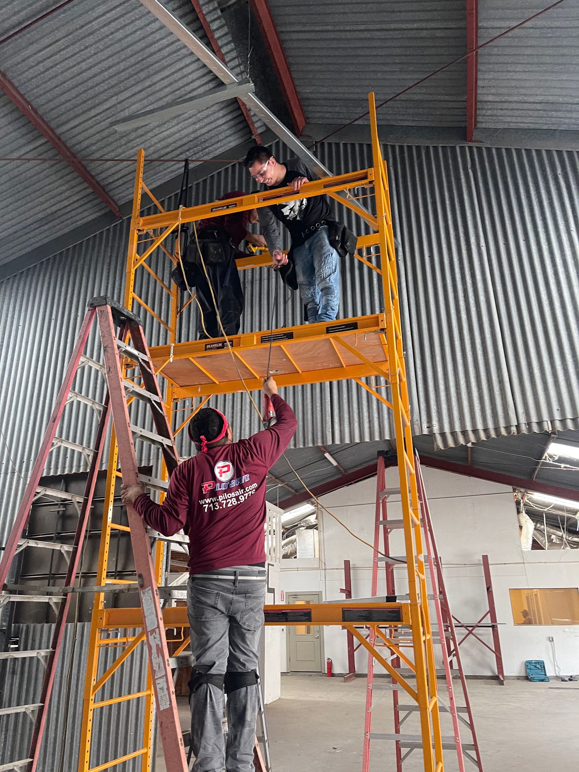 Three workers on a yellow scaffolding inside a corrugated metal building; one person on a ladder below.