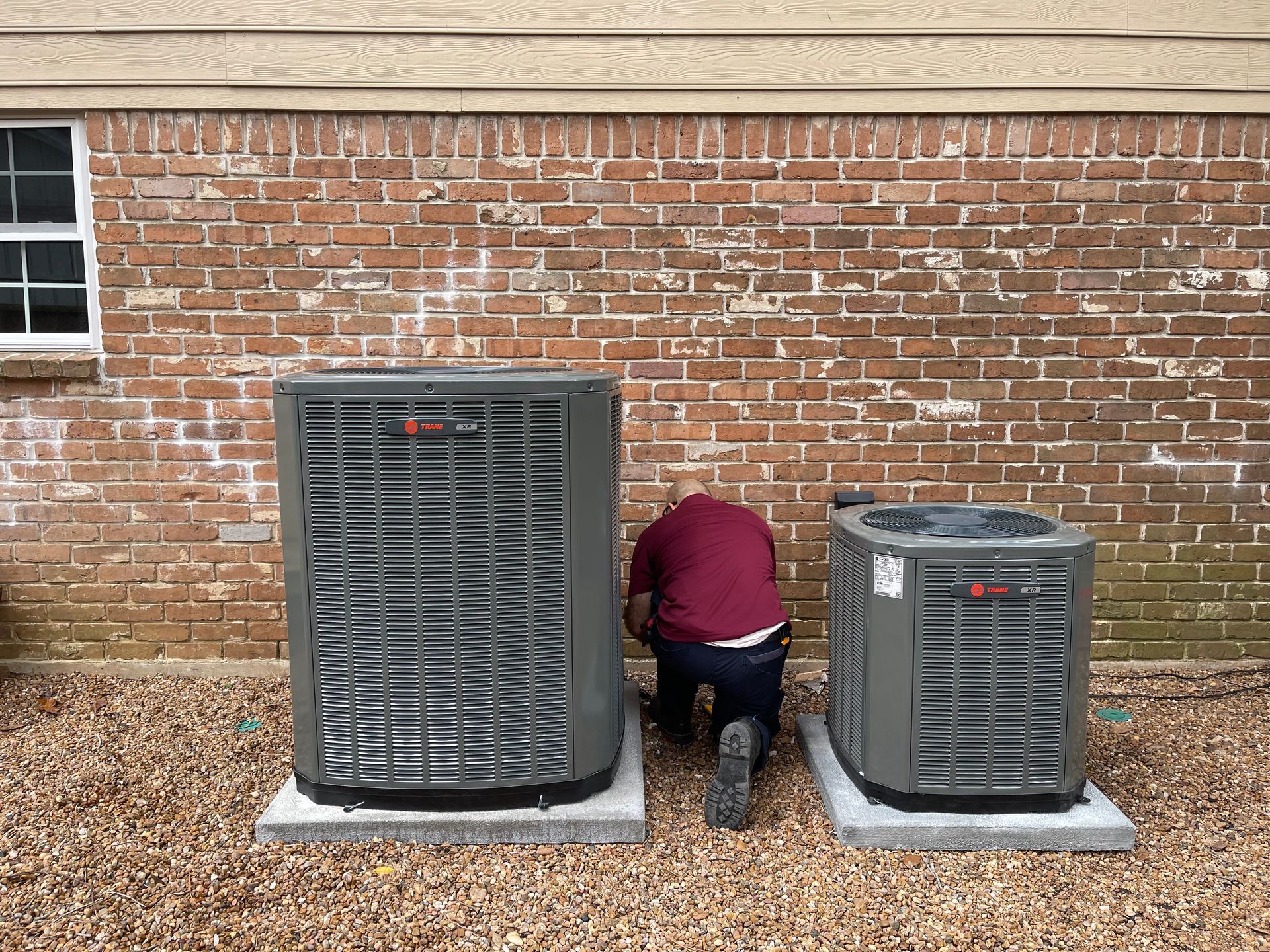 A person kneels beside two gray HVAC units on concrete pads, against a brick wall.
