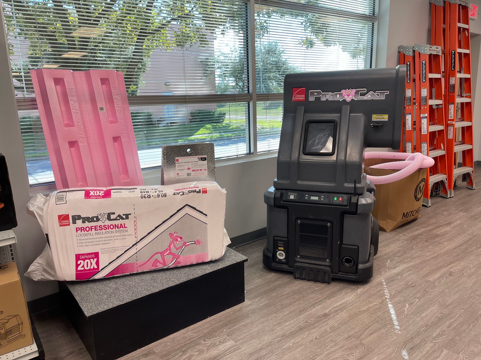 Construction supplies near a window: Pink insulation, pallet, Pro-Cote machine, ladders, and a cardboard box.
