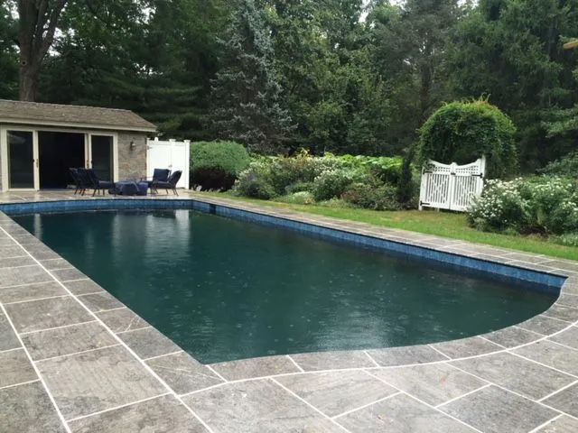 Rectangular pool with dark water and stone patio, small building, and landscaping in a backyard.