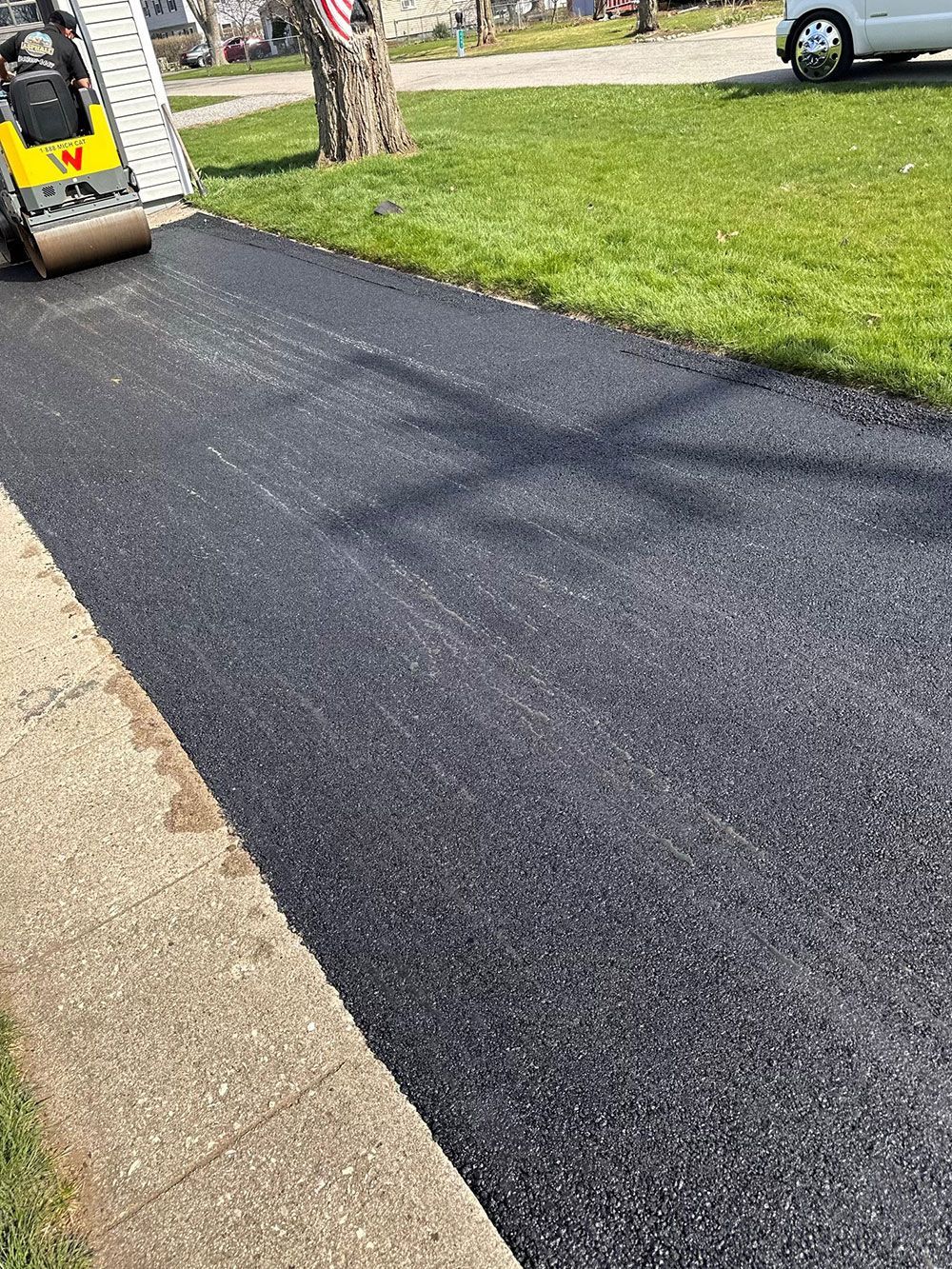 Asphalt driveway being rolled by a small construction machine, beside a green lawn and concrete curb.