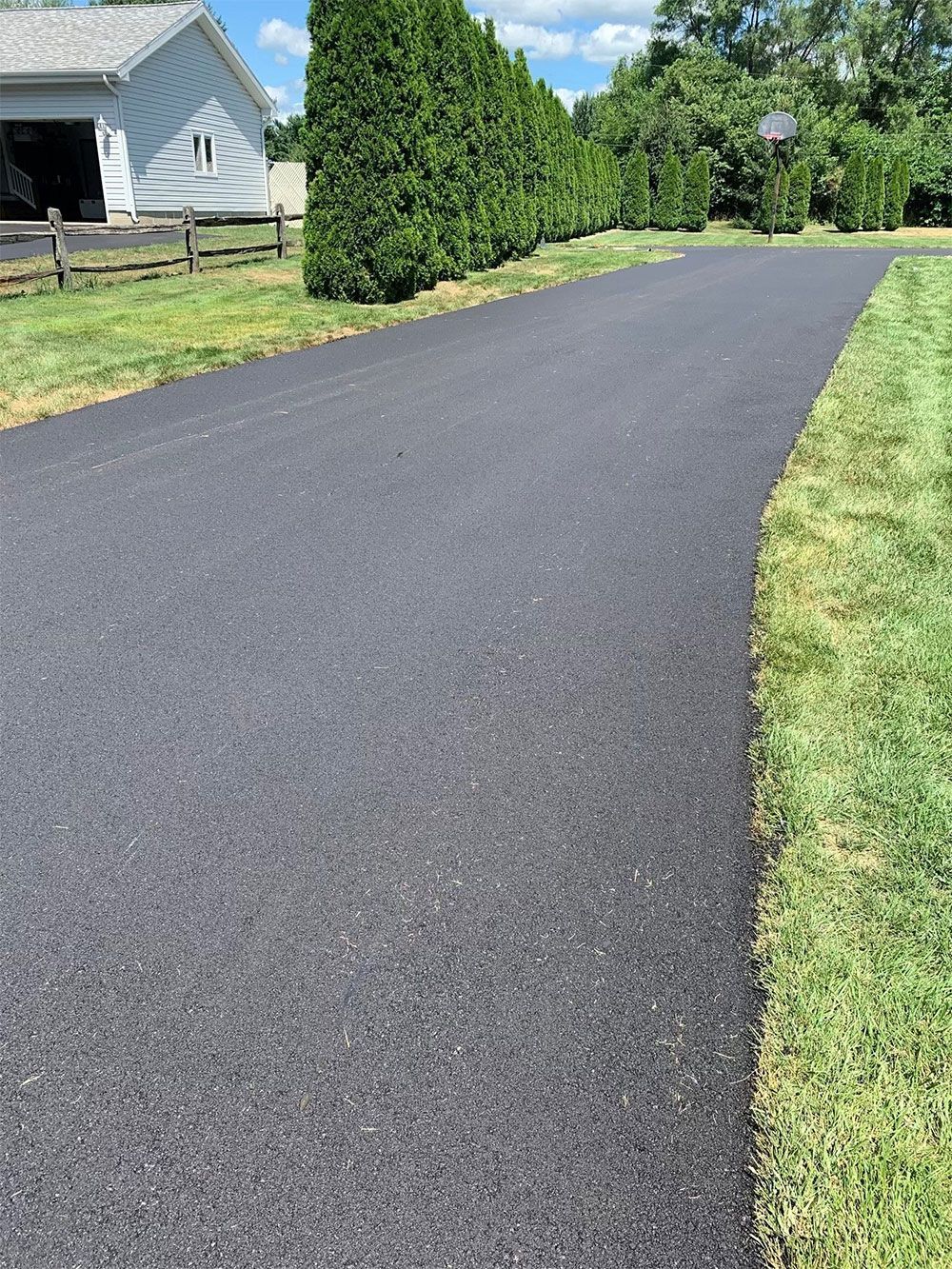 Asphalt driveway bordered by green grass and trees, leading toward a house.