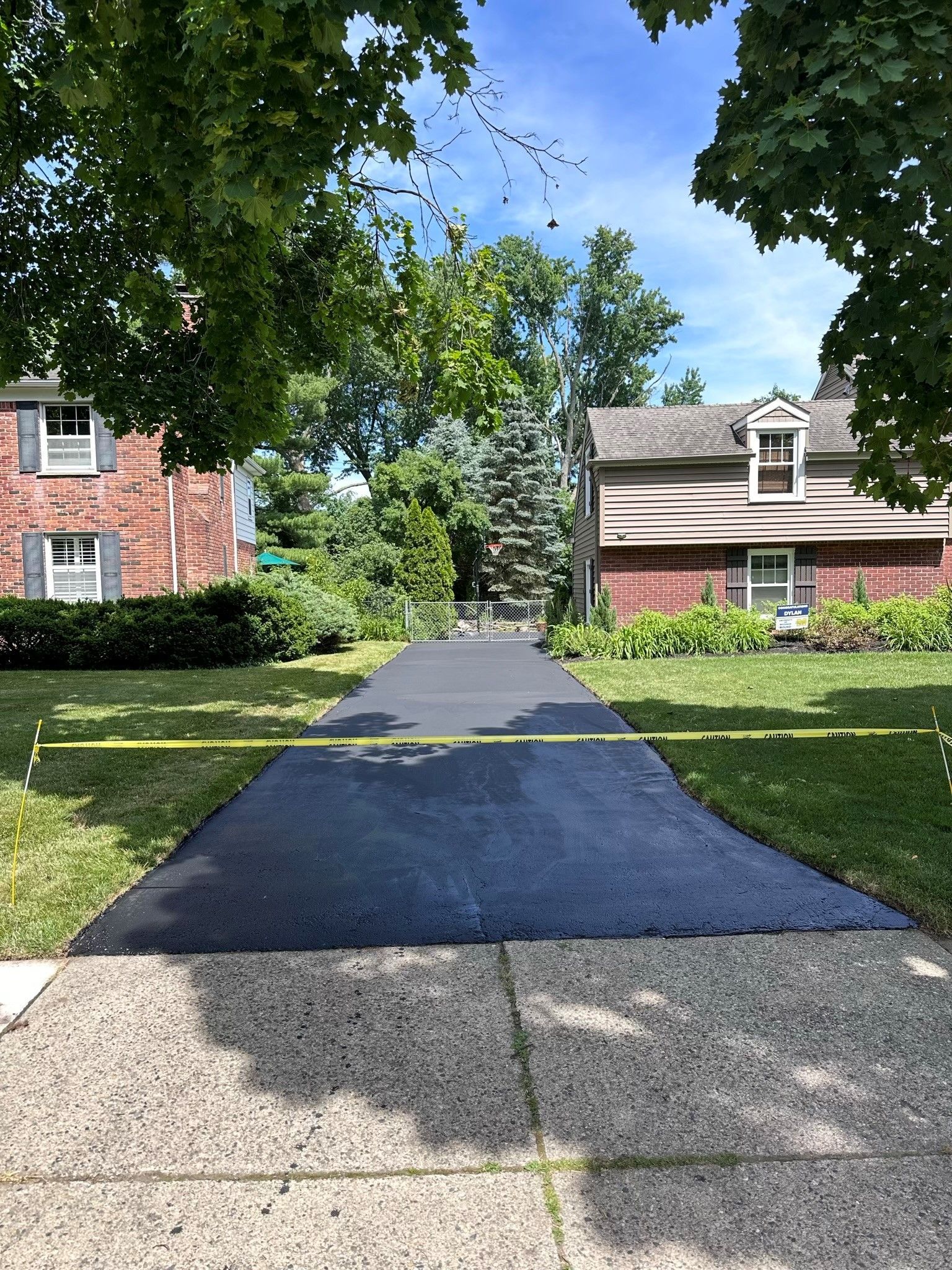 Newly paved driveway with yellow caution tape, between two houses with green lawns and trees.