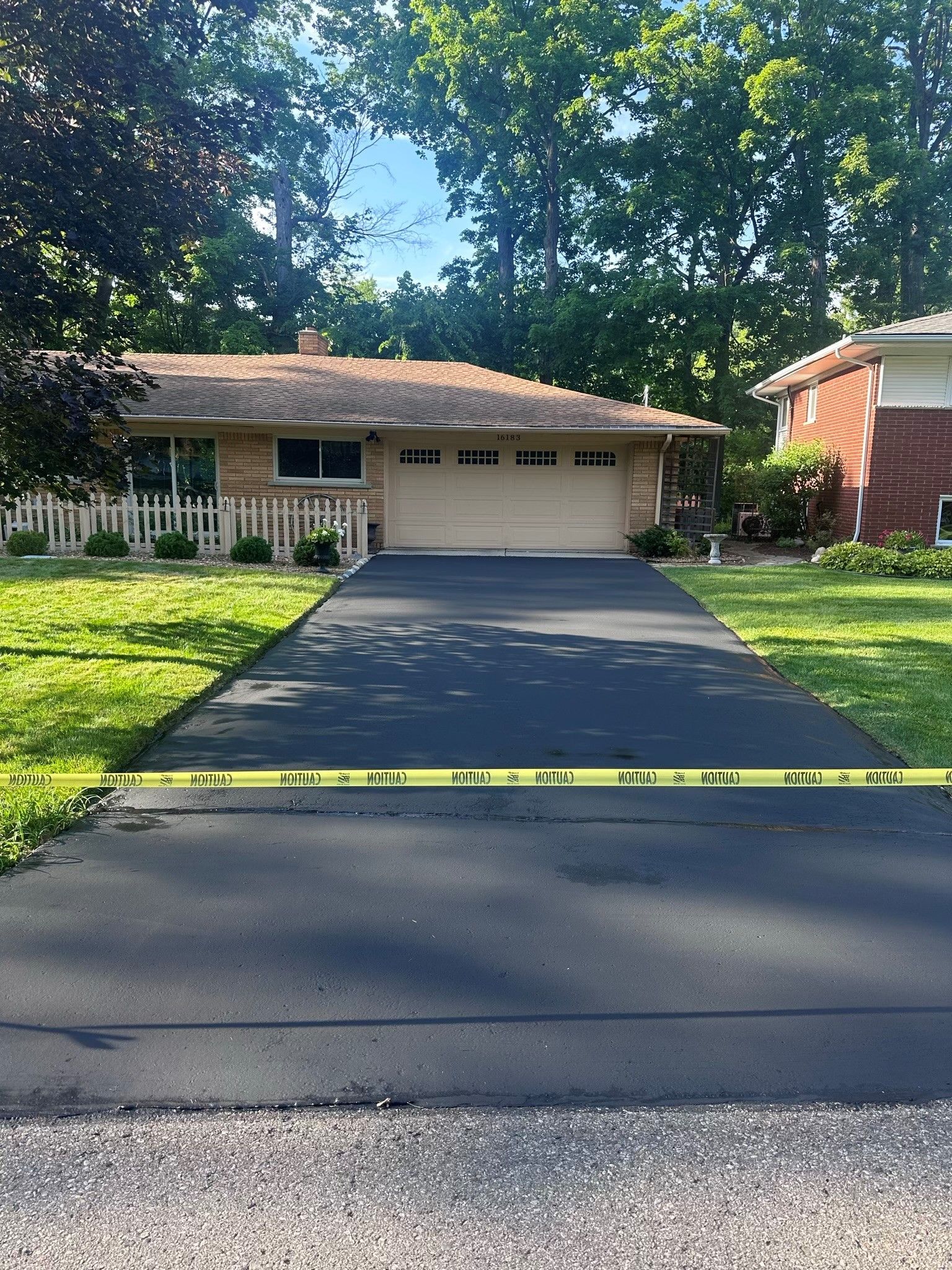 A single-story brick house with a newly paved driveway, yellow caution tape across the pavement.