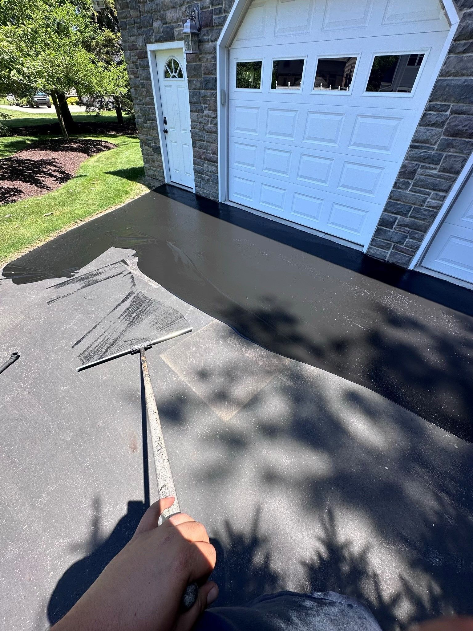 Person sealing a freshly paved black driveway with a squeegee near a white garage door.