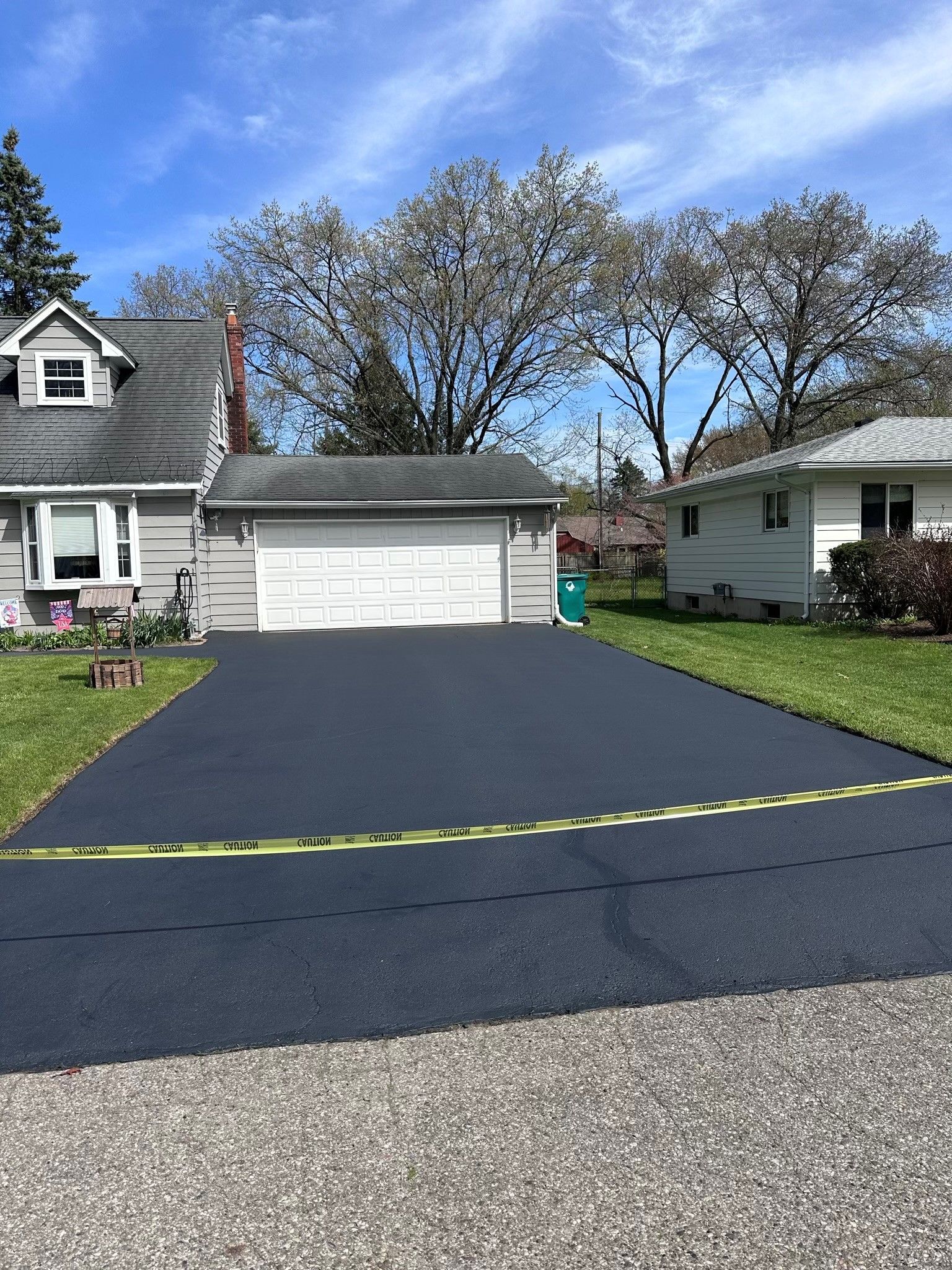 Freshly paved black driveway with caution tape, houses, and trees on a sunny day.