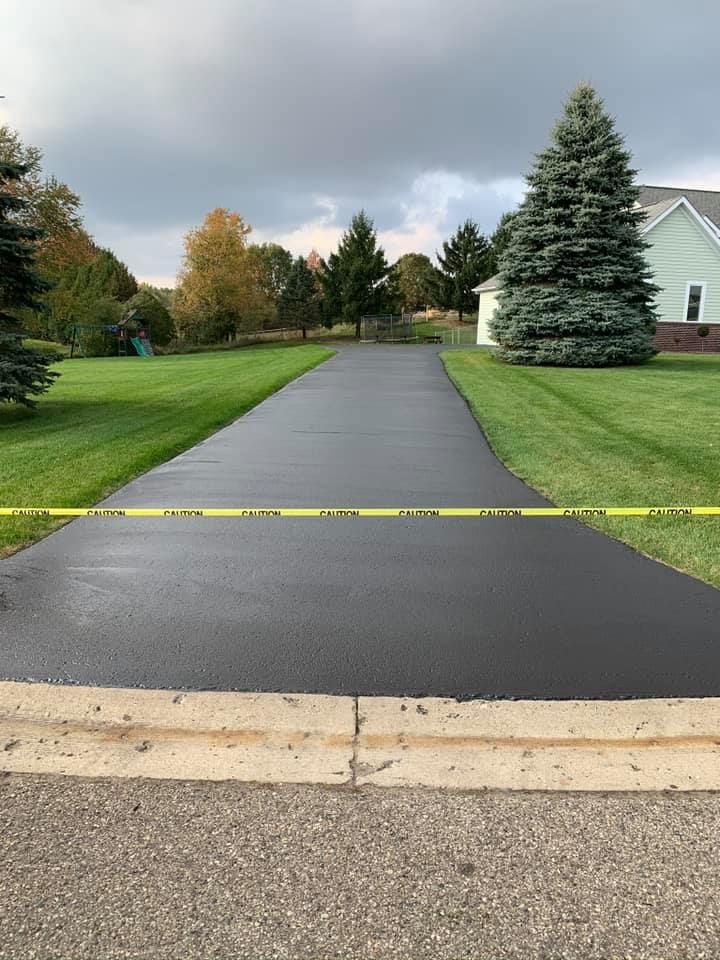 Freshly paved black driveway with yellow caution tape, flanked by green grass and trees.