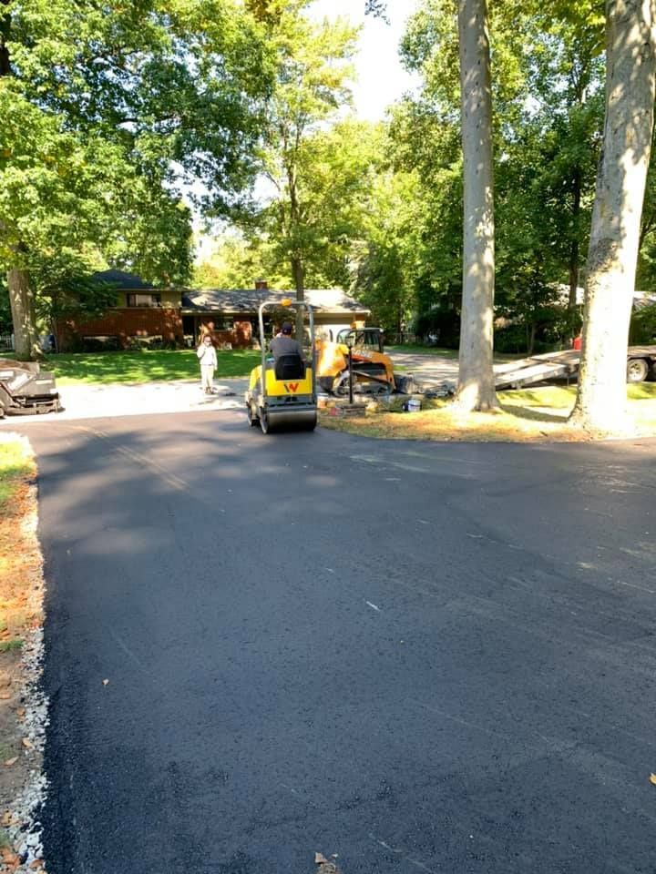Asphalt driveway being compacted by a small roller; person operating the machinery; trees in the background.