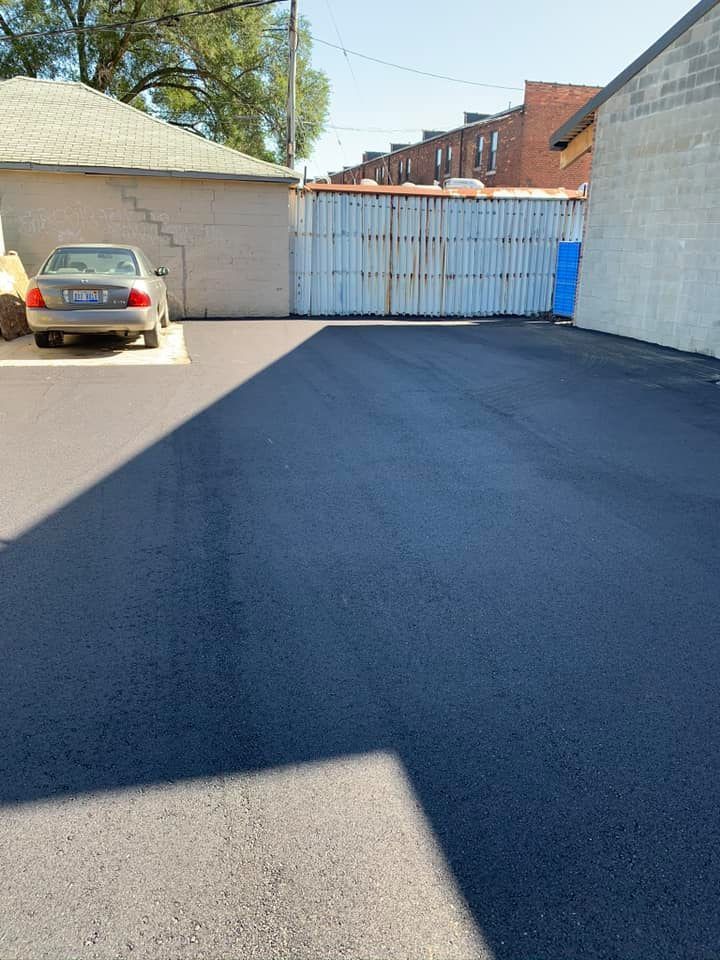 Paved parking area; gray car, corrugated metal gate, brick buildings in background.
