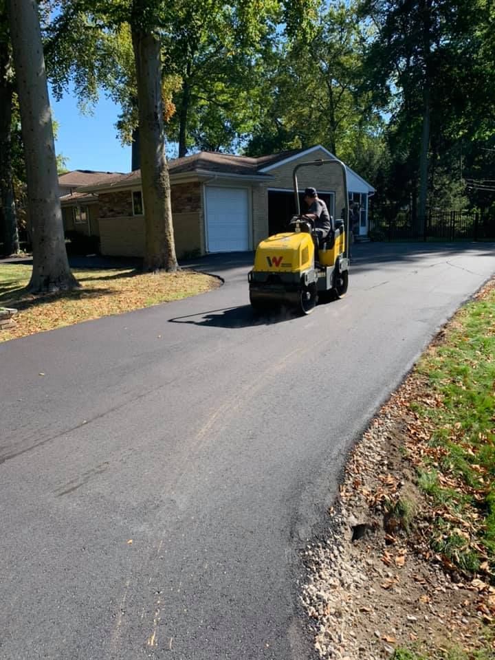 Person operating a yellow asphalt compactor on a newly paved driveway, next to a house and trees.