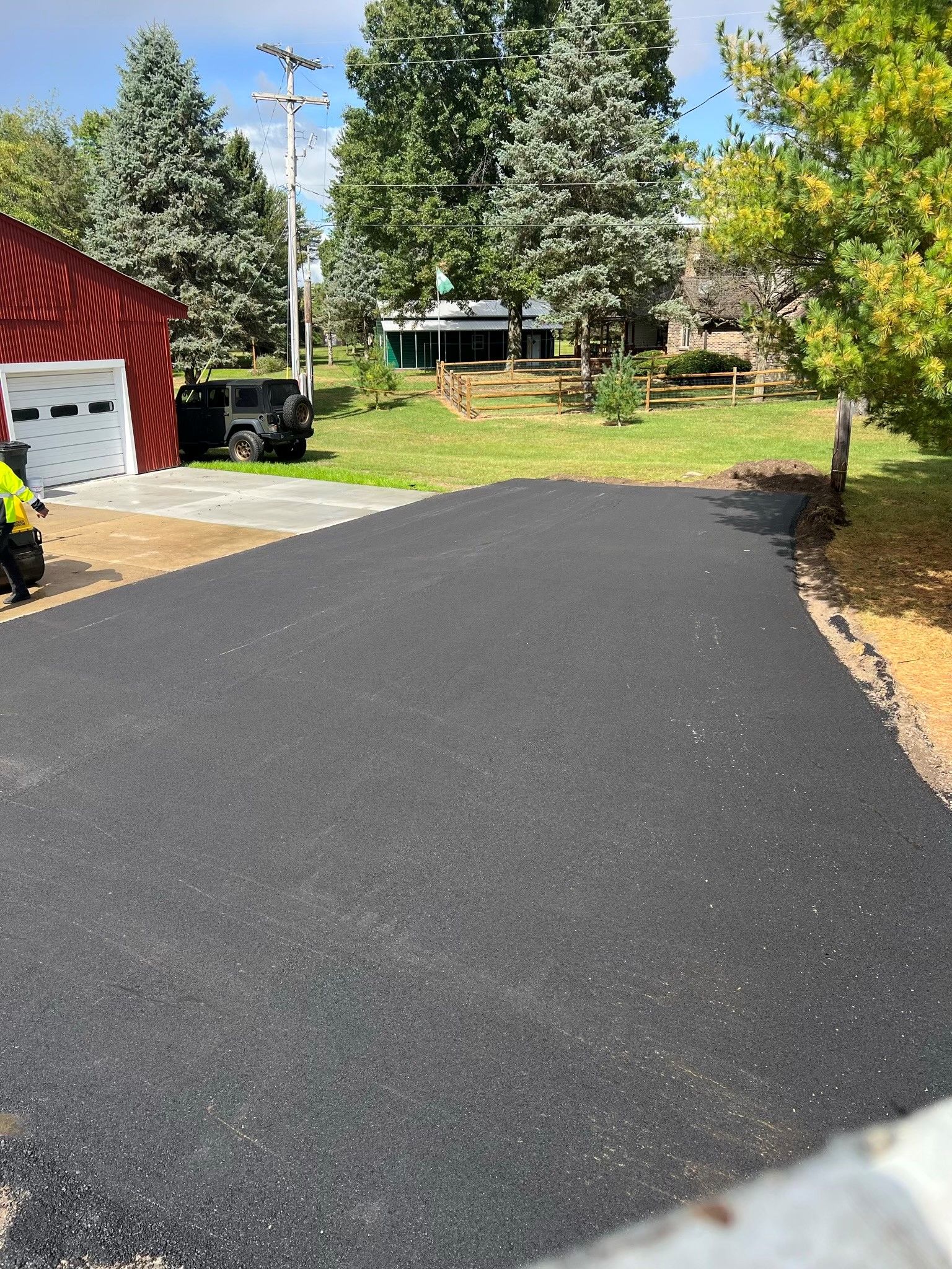 Newly paved black asphalt driveway leading towards a red building and grassy area under a blue sky.