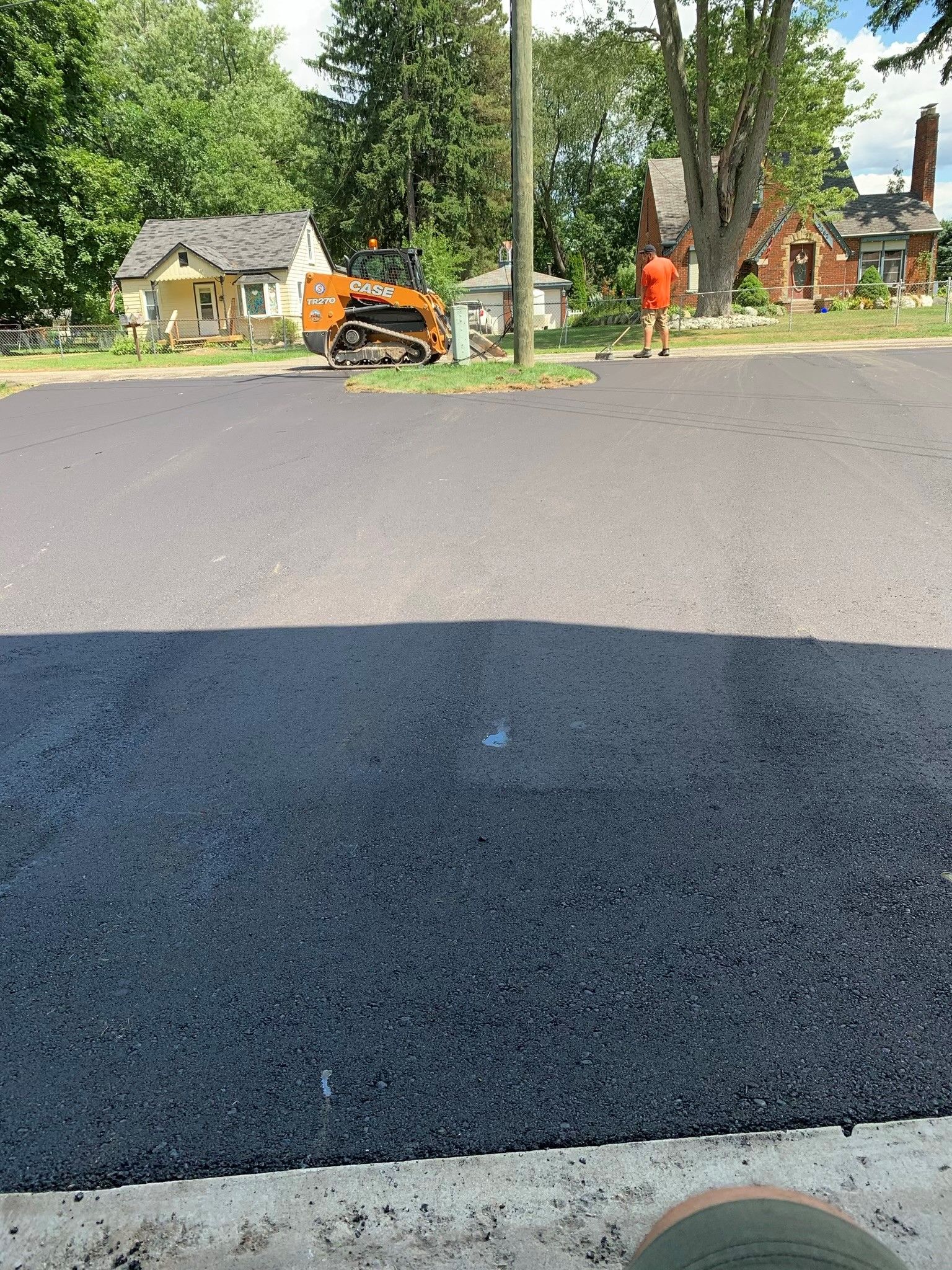 Freshly paved asphalt road; construction worker raking; small machinery; houses in background.