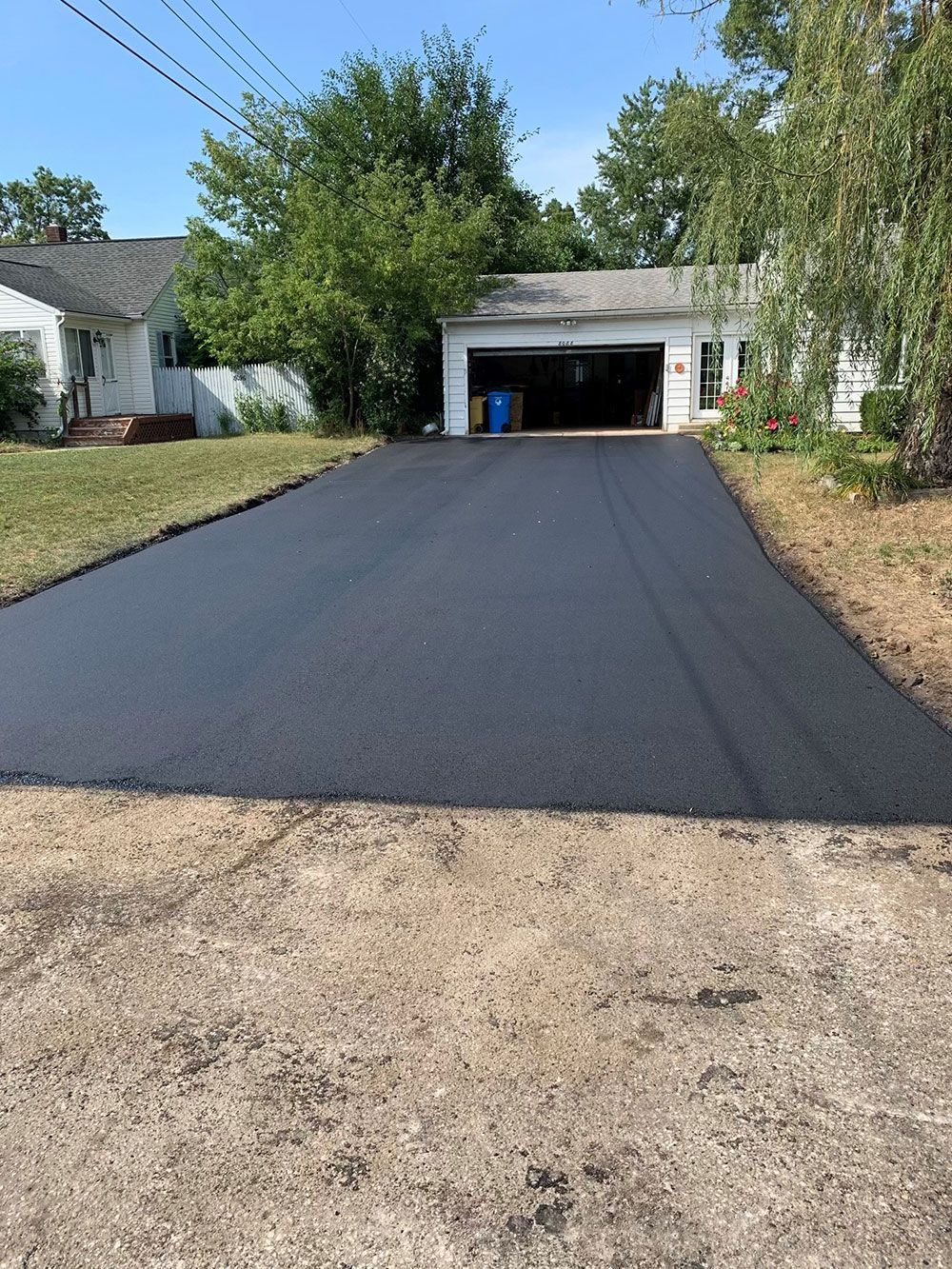 Newly paved asphalt driveway leading to a garage door, with a house and trees in the background.