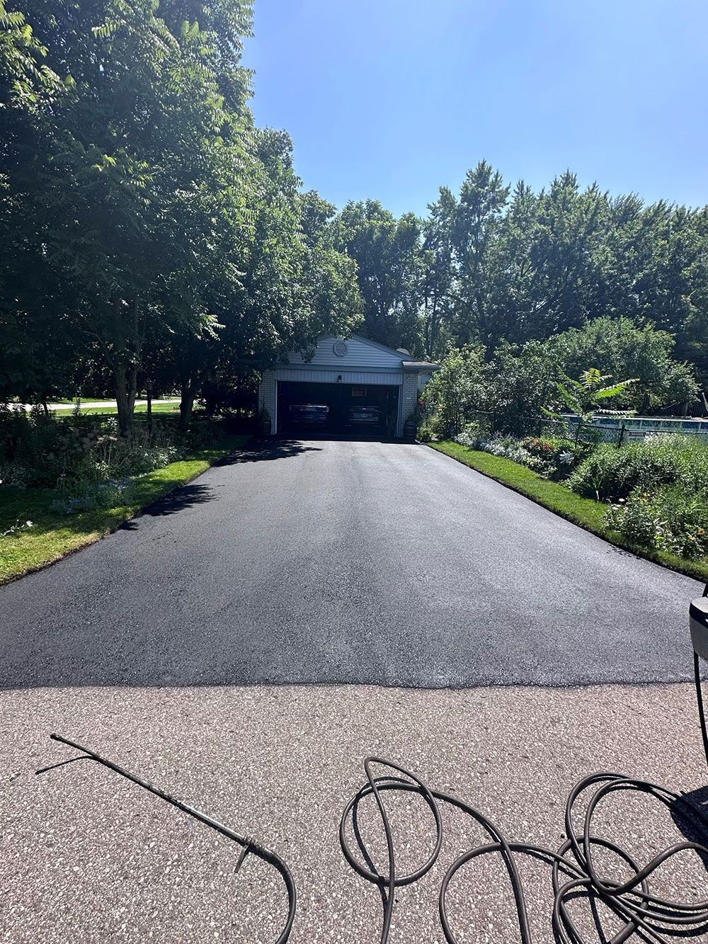 Newly paved asphalt driveway leading to a garage, surrounded by green trees and grass on a sunny day.