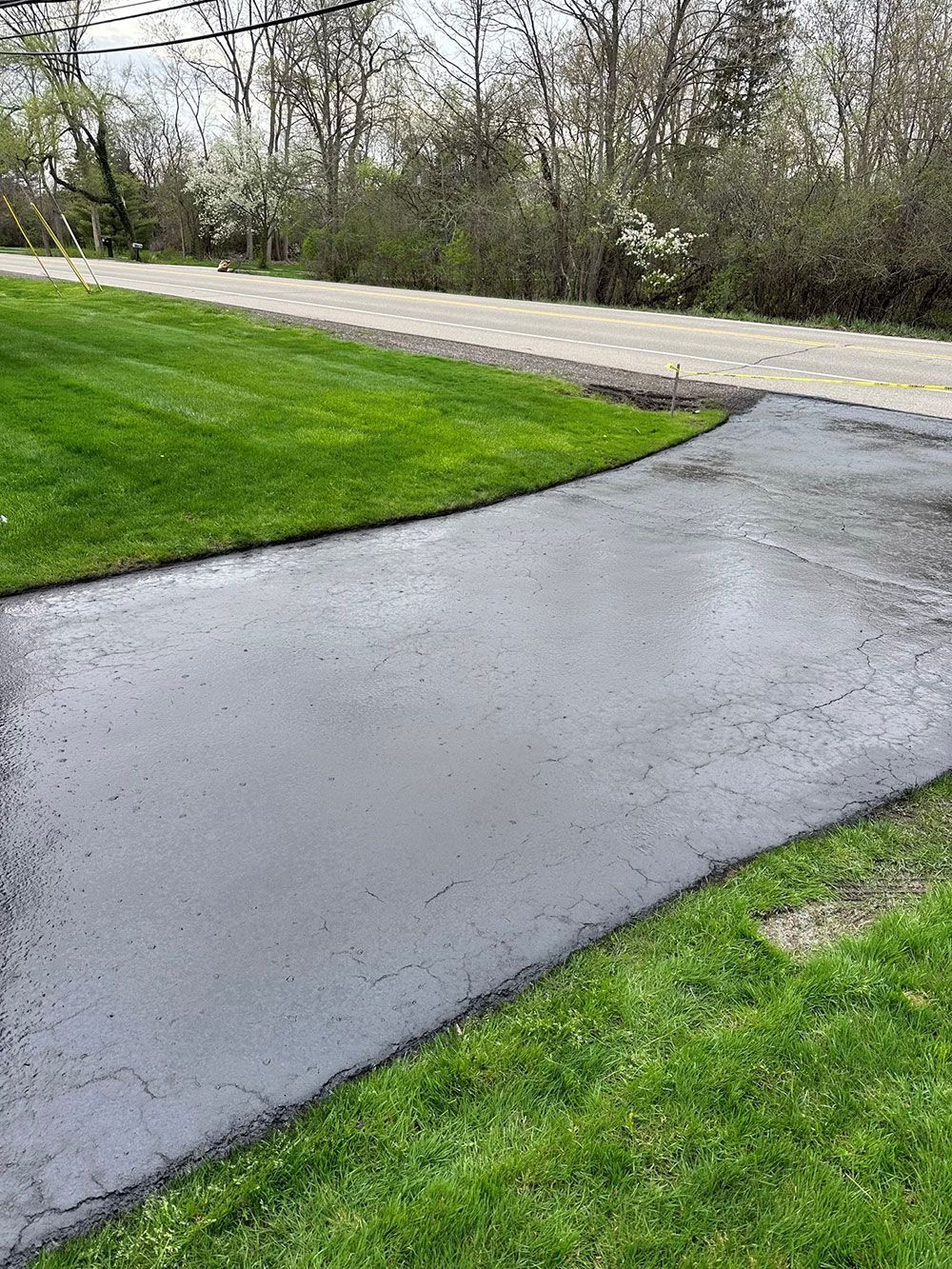 Black asphalt driveway next to a green lawn, leading to a road with trees in the background.