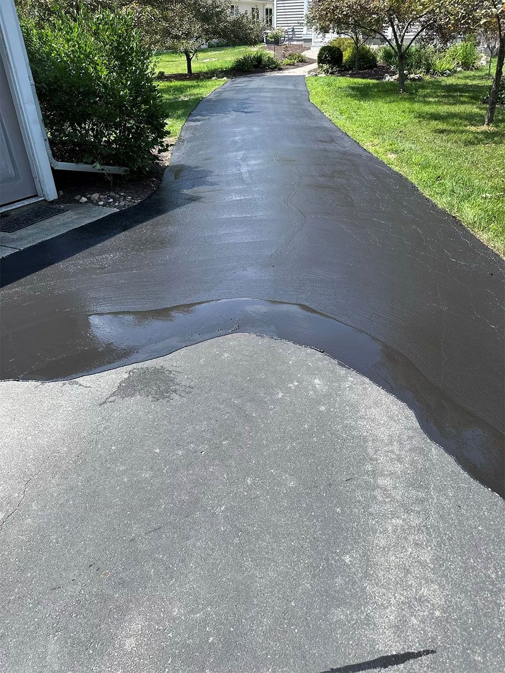 Paved driveway with a water puddle in the center, flanked by green grass and a house.
