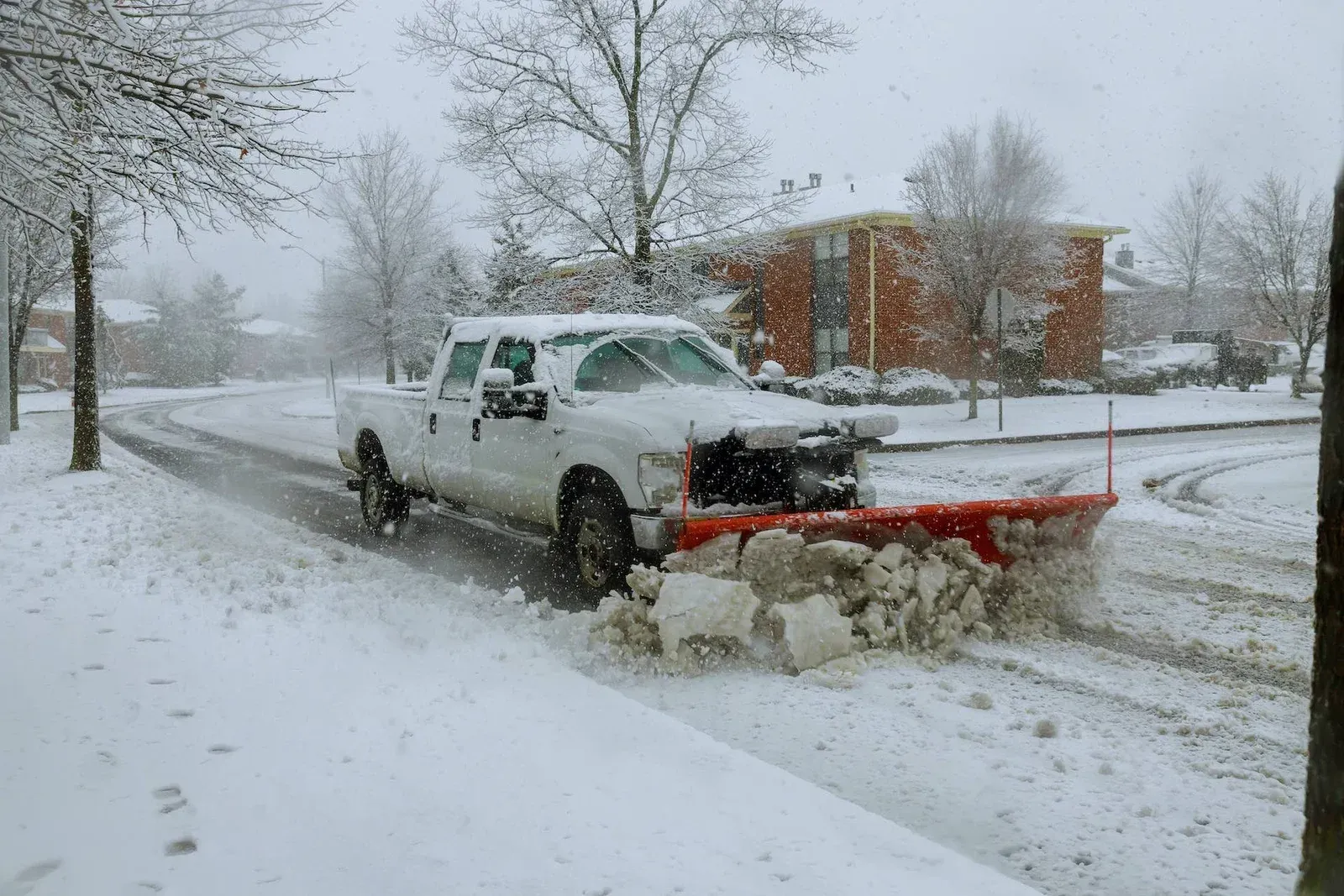 White pickup plowing snow on a city street, pushing snow toward the curb in a winter storm.