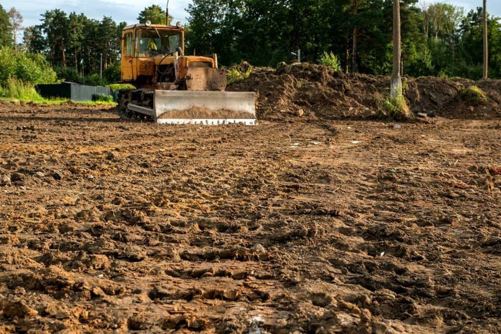 Bulldozer clearing a muddy field with dirt piles and trees in the background