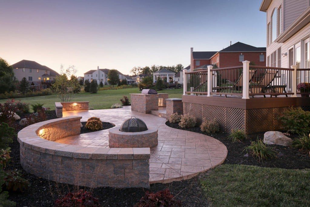 Curved stone fire pit and patio in a landscaped backyard at dusk, beside a house with outdoor seating