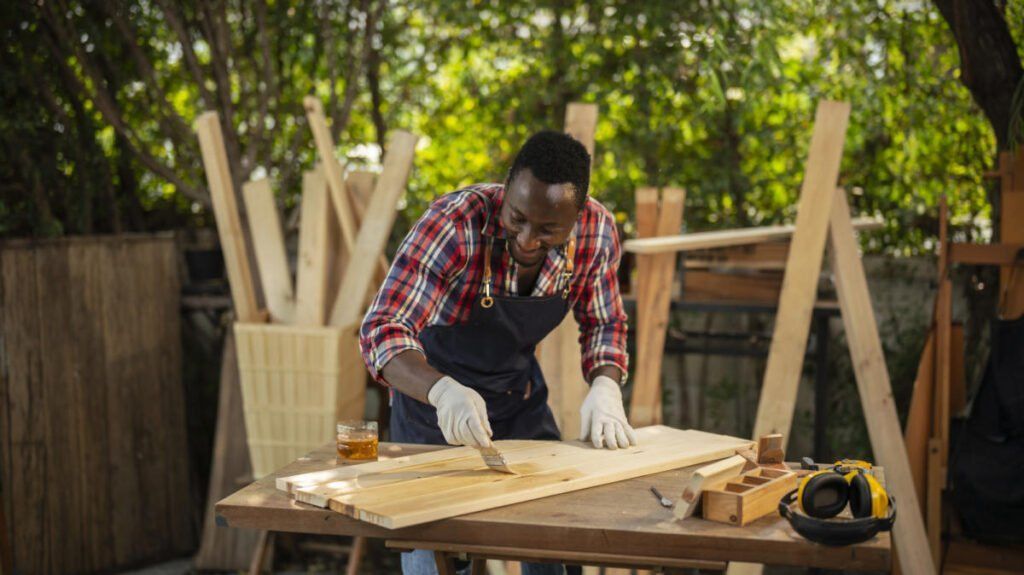 Person building a wooden chair at an outdoor workbench, wearing gloves and a red plaid shirt