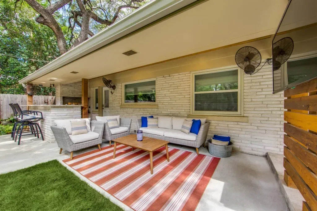 Covered patio with striped rug, outdoor seating, coffee table, and wall-mounted fans beside a brick house