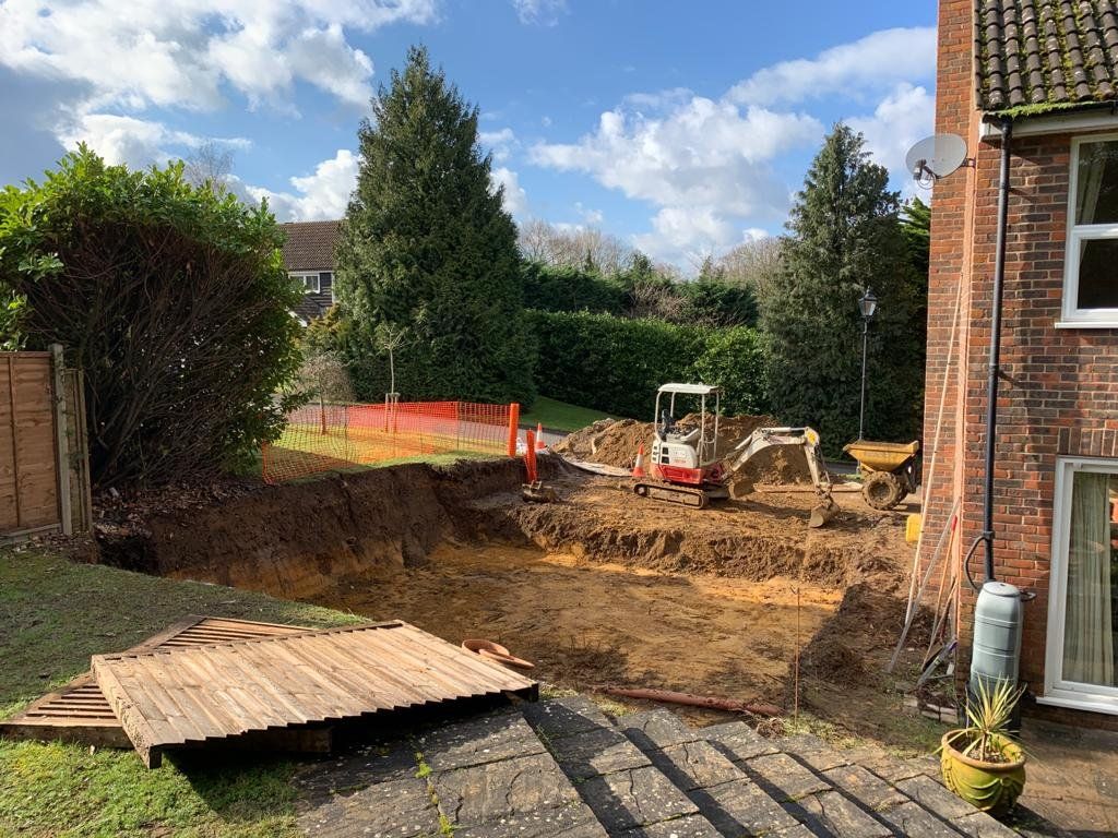 Backyard excavation beside a brick house, with dirt piles, a mini digger, and a wooden pallet in the foreground