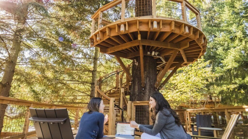 Two people sit at a picnic table beneath a wooden treehouse deck in a forest setting.