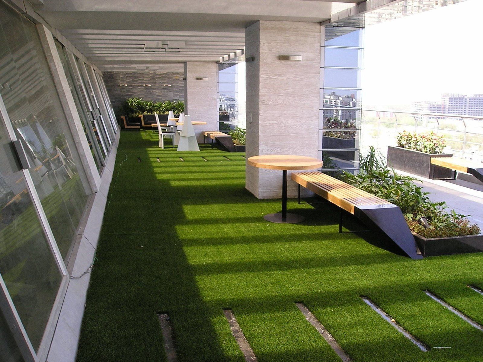 Modern patio with green turf, white columns, benches, and potted plants in bright sunlight