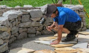 Worker in blue shirt laying stone pavers on a garden walkway beside a stacked stone wall