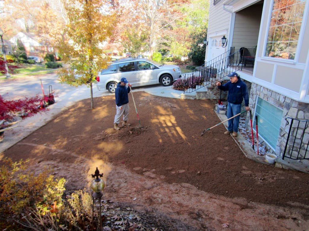 Two workers raking leaves in a driveway beside a house, with autumn trees and a parked white car.