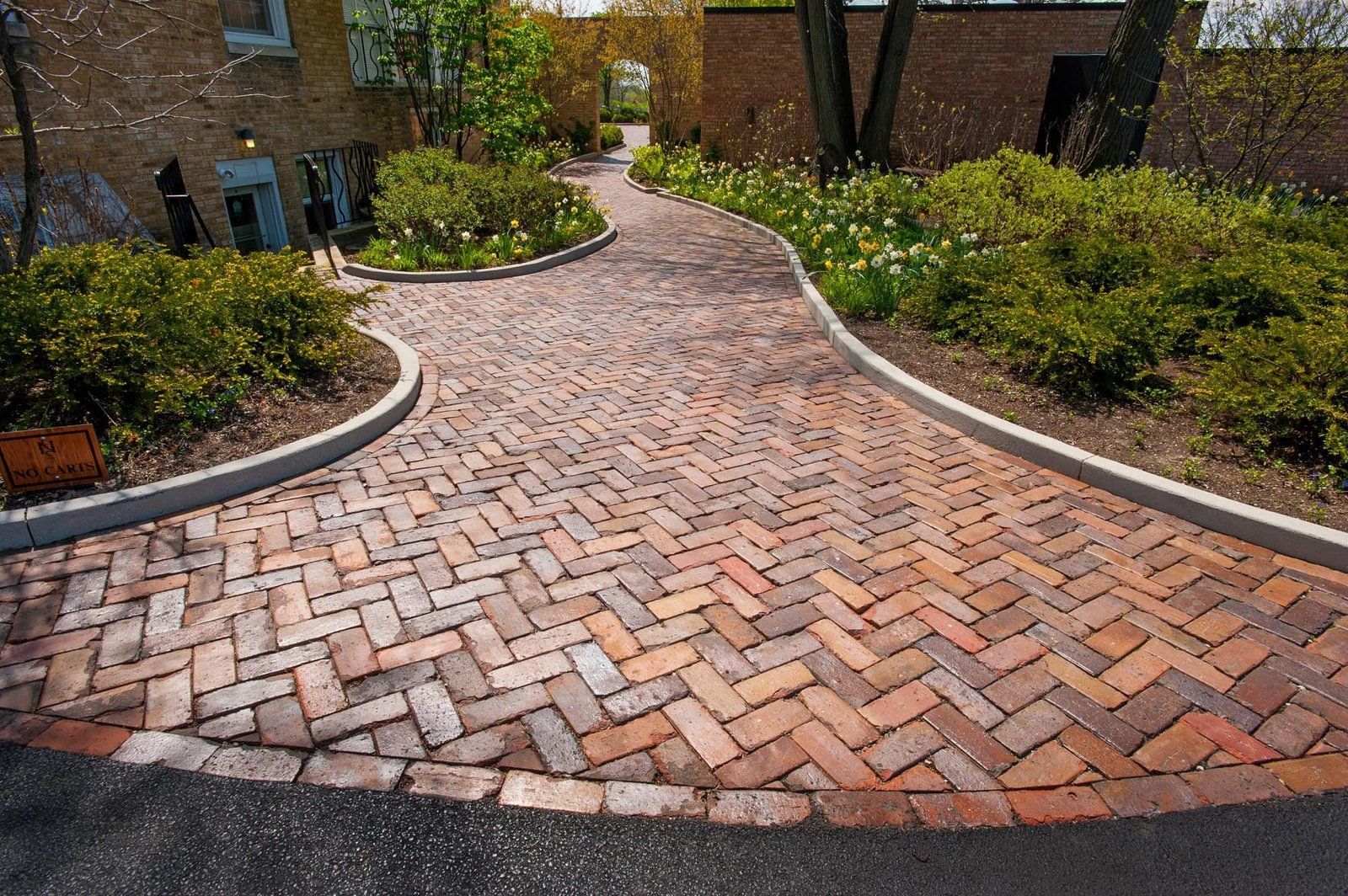 Brick-paved garden walkway curving between landscaped shrubs and brick buildings